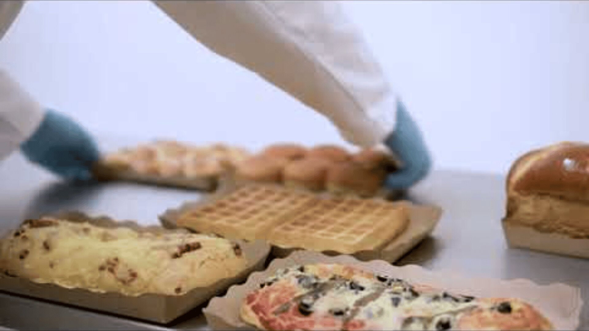 A person wearing a white coat and blue gloves arranges baked goods, including waffles, bread, and pastries with various toppings, on a metal surface.