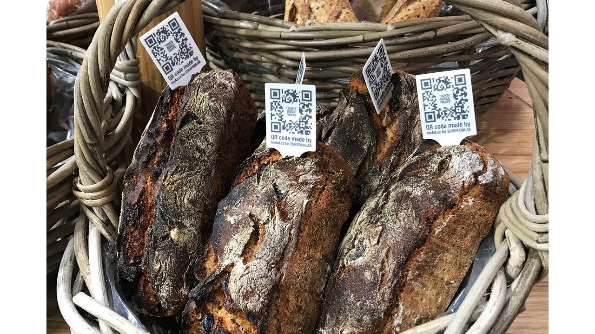 A basket of rustic loaves of bread with dark, crusty tops. Each loaf has a small sign with a QR code attached, and more bread is visible in baskets in the background.