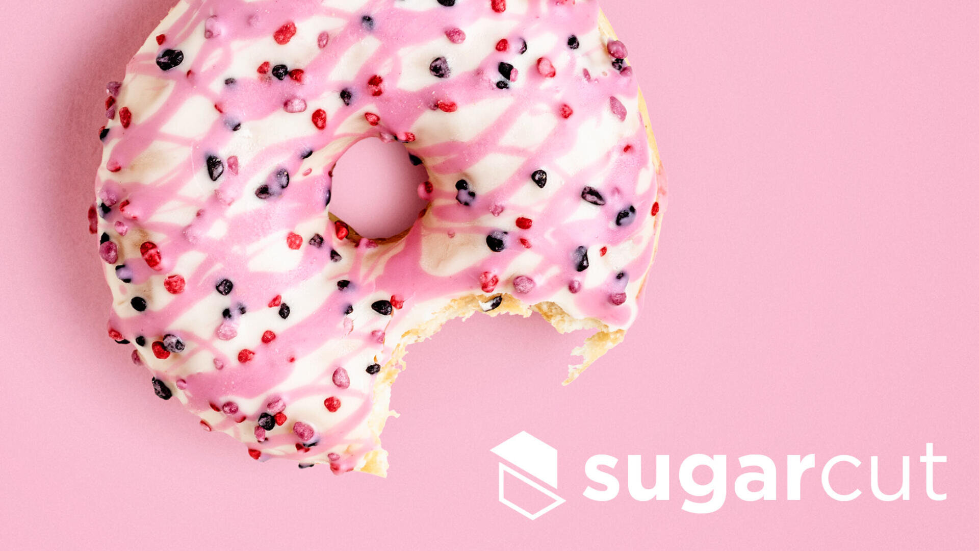 Ein Donut mit rosa-weißer Glasur, bestreut mit kleinen roten und schwarzen Bonbonstückchen, wird angebissen. Der Hintergrund ist rosa, und in der unteren rechten Ecke erscheint das Sugarcut-Logo.