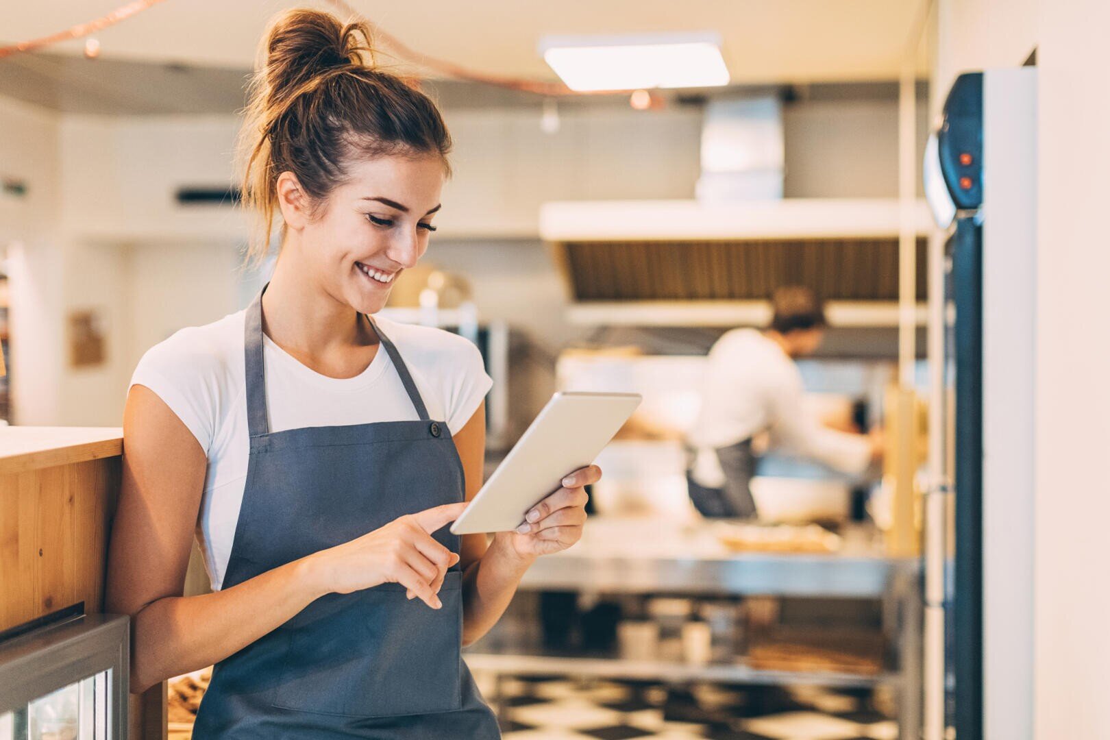 Eine lächelnde Frau in einer grauen Schürze benutzt ein Tablet in einer hellen, modernen Küche. Eine andere Person arbeitet im Hintergrund in der Nähe von Öfen und Kochgeräten.