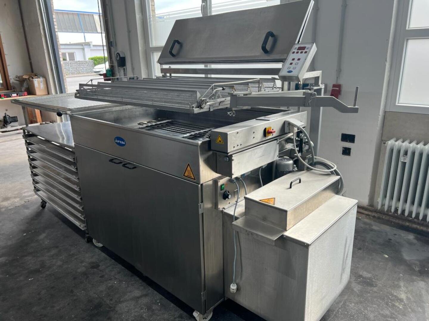 Eine große Industriemaschine aus rostfreiem Stahl mit Bedienfeld und offenem Deckel, die in einer Werkstatt mit hellen Fenstern und Betonboden steht. An der Maschine sind Warnschilder und Anschlusskabel sichtbar.