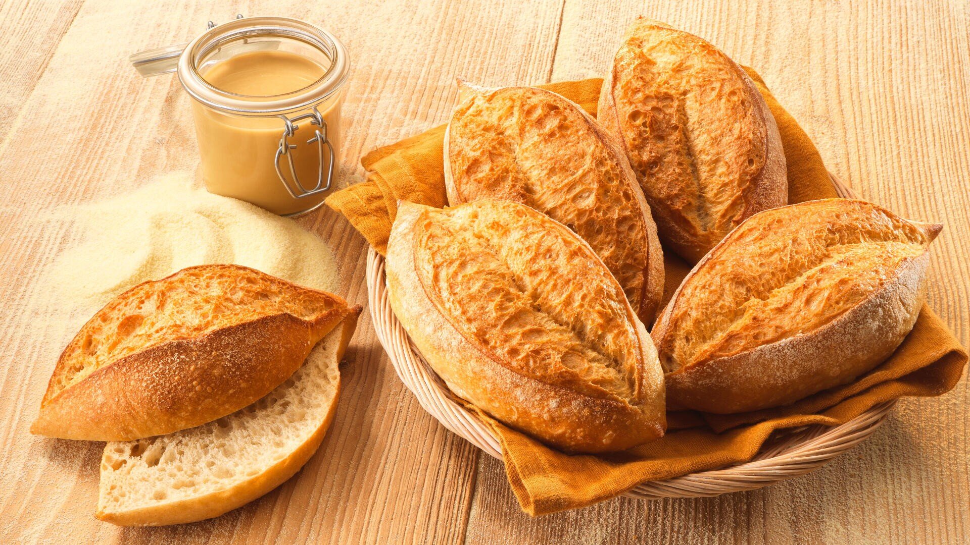 Ein Korb mit fünf knusprigen Brötchen auf einer Stoffserviette steht auf einem Holztisch neben einem halbierten Brötchen, einem Glas Brotaufstrich und einem kleinen Haufen Mehl oder Grieß.