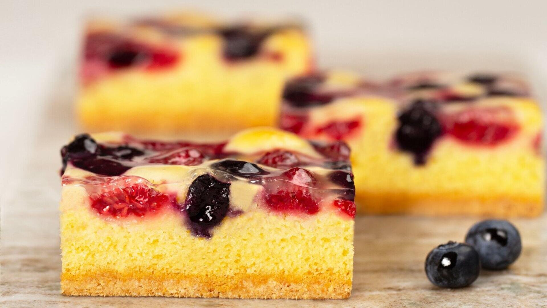 Close-up of three slices of sponge cake topped with a clear glaze, raspberries, and blueberries. Two fresh blueberries are placed next to the cake on a light surface.