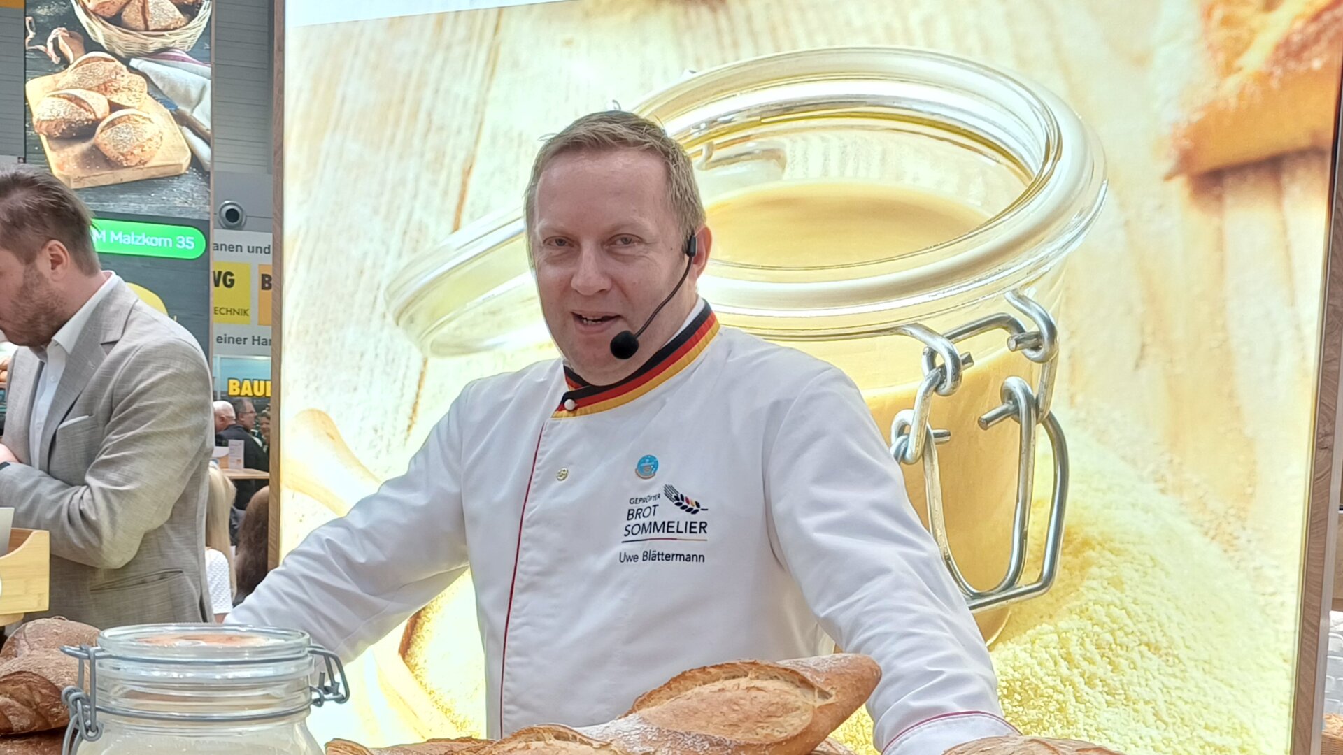 Ein Bäcker steht hinter einem Tisch und präsentiert verschiedene Brote und Gebäcke, ein großes Glas und kleine Gläser. Daneben steht eine Meisterurkunde, und im Hintergrund unterhalten sich Menschen auf einer Messe oder Ausstellung.