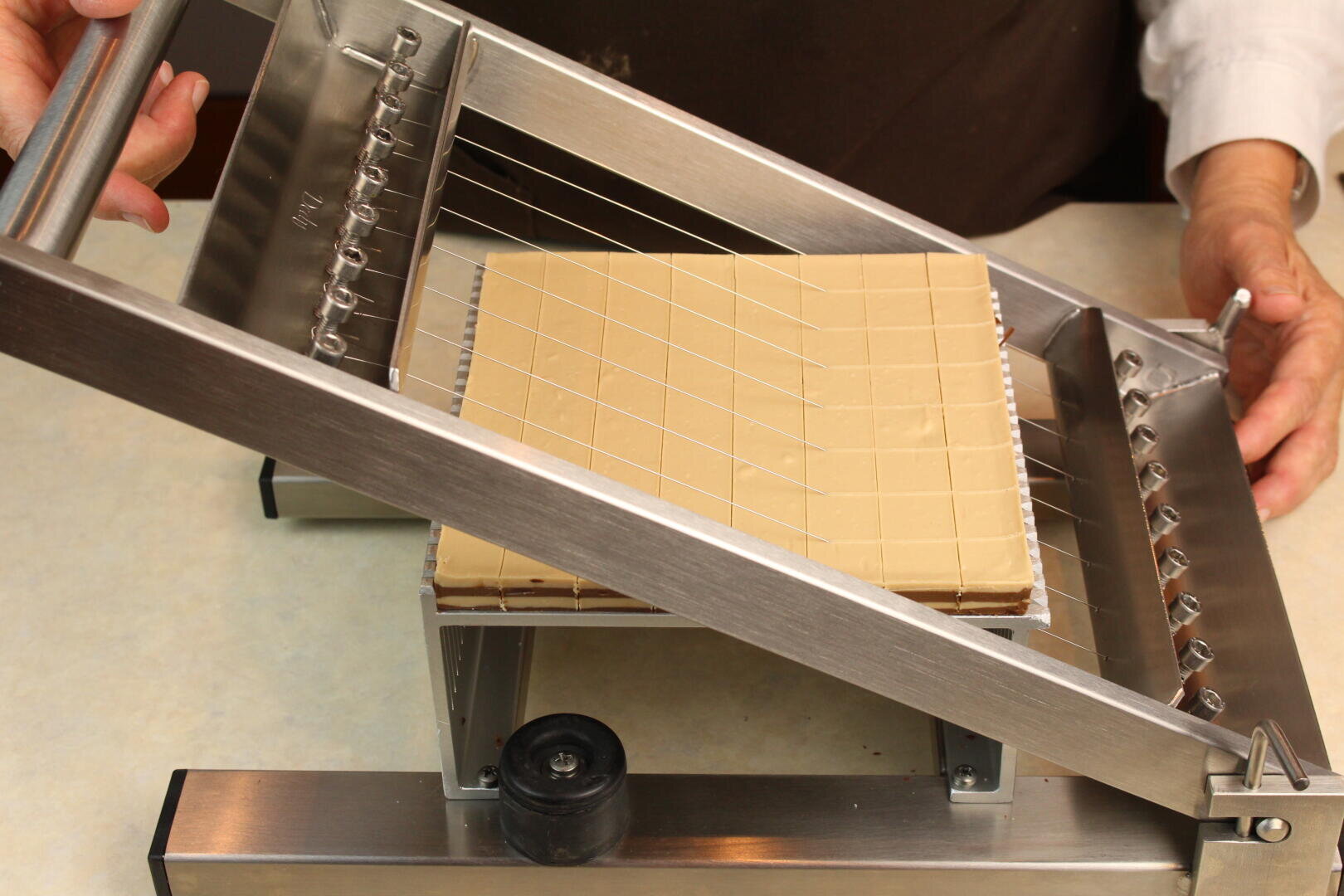 Eine Person schneidet mit einem Drahtschneider aus rostfreiem Stahl einen großen quadratischen Block aus beigem Fondant oder Nougat auf einer Arbeitsplatte in kleinere Würfel.