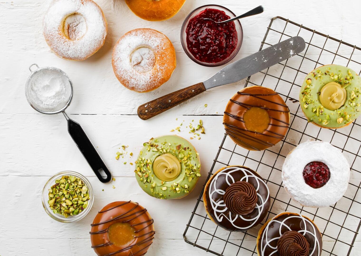 An assortment of donuts with various toppings, including powdered sugar, pistachios, chocolate, and fruit jam, arranged on a white table with a wire rack, knife, and bowls of jam and pistachios.
