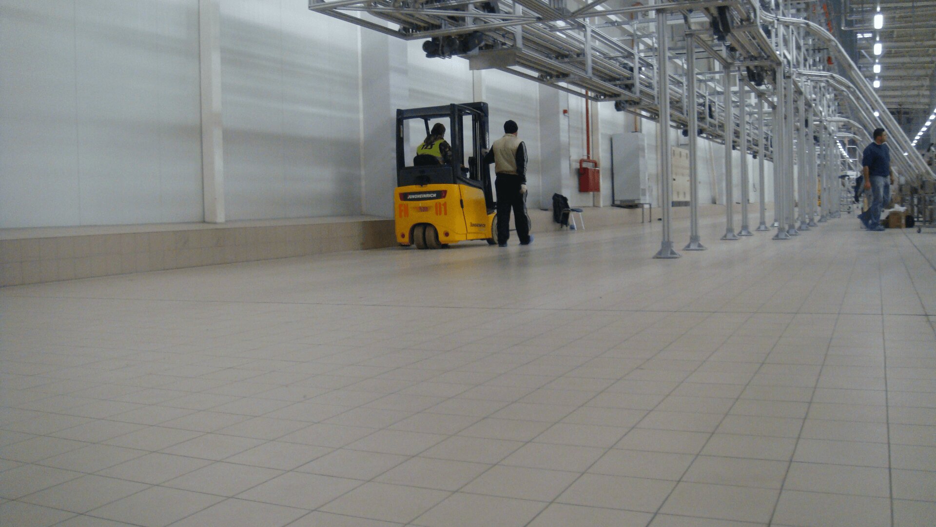 A yellow forklift operated by a worker moves down a tiled factory floor, while another person walks beside it. Metal pipes and supports run along the right side of the spacious, well-lit industrial area.