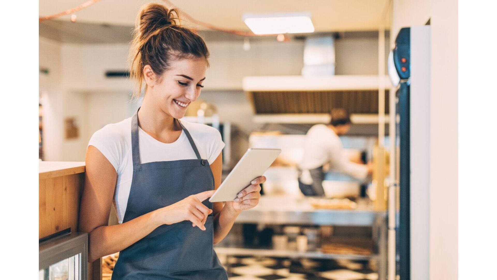 A smiling woman wearing a gray apron uses a tablet in a bright kitchen, with another person working in the background.