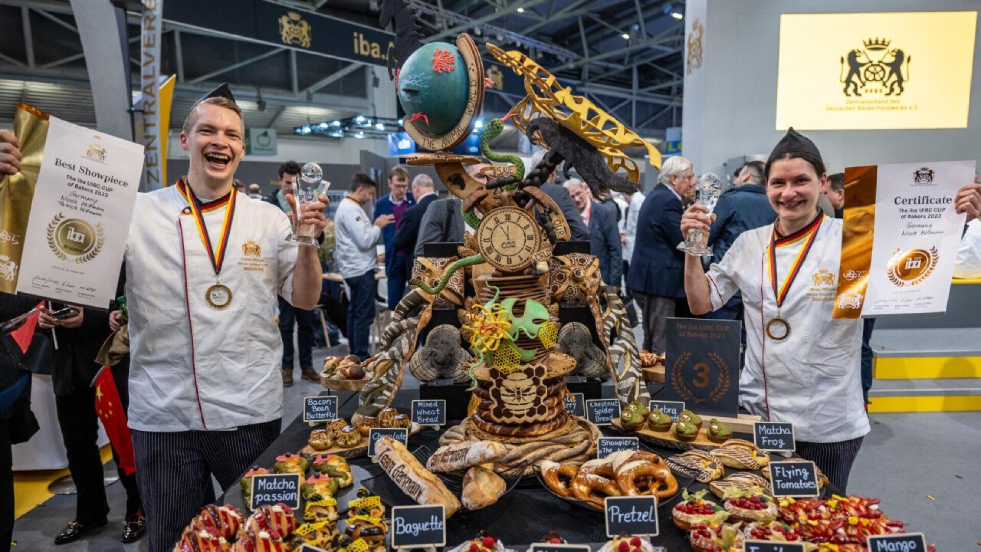 Two smiling chefs hold awards beside an elaborate display of pastries, breads, and desserts at a baking competition. The colorful table centerpiece includes animal and clock-themed decorations. People observe in the background.
