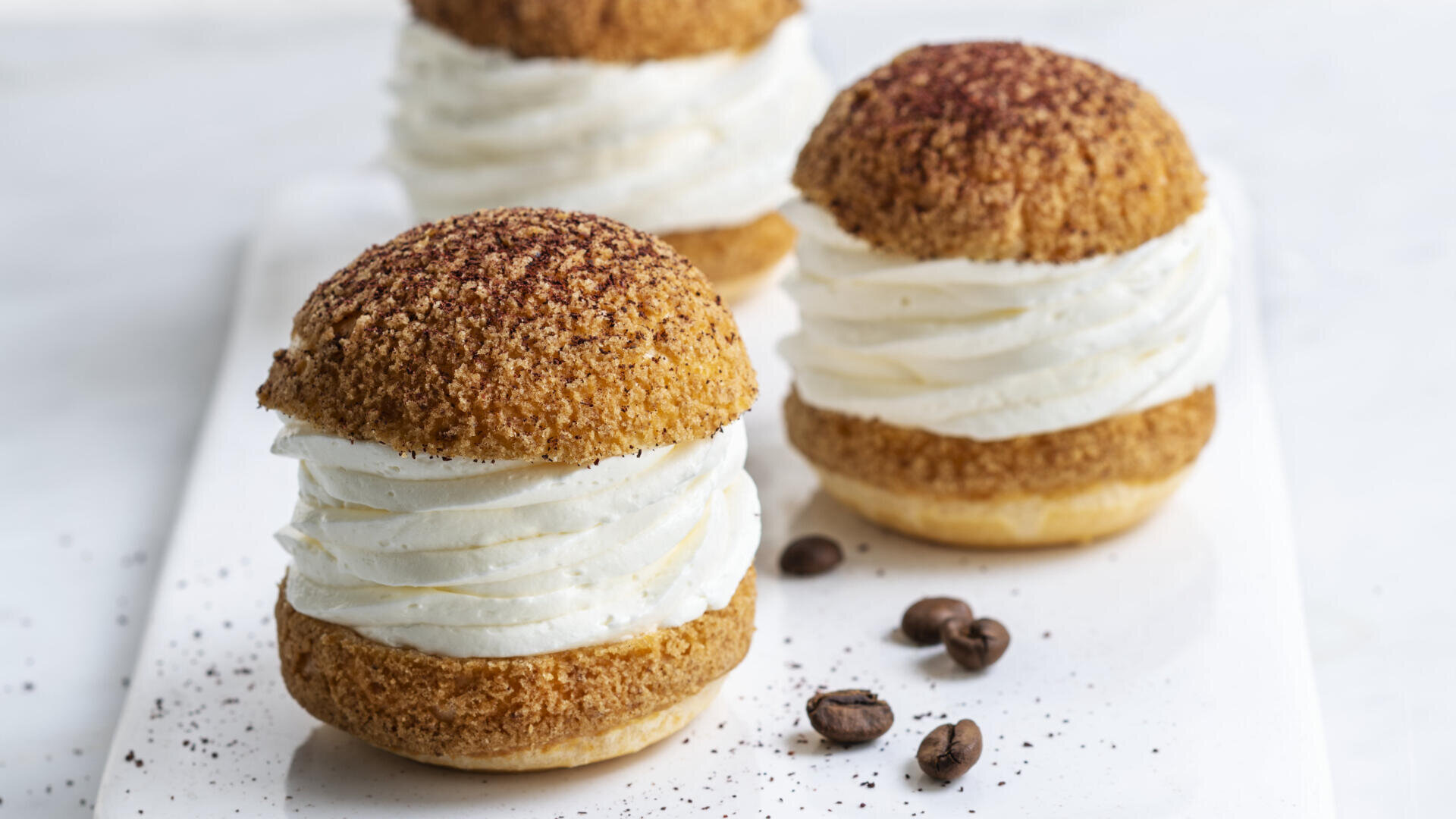 Three cream-filled choux pastries with crisp, golden tops are arranged on a white surface, surrounded by a few coffee beans and scattered coffee powder.