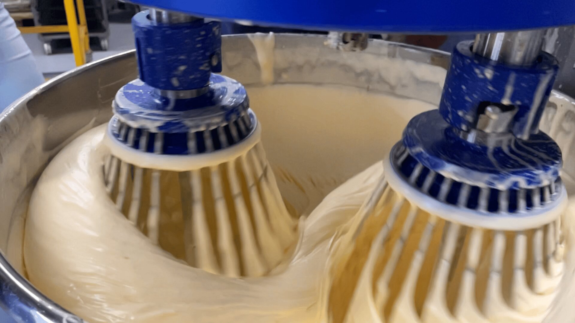 Close-up of two large industrial mixers blending thick, creamy batter in a stainless steel bowl, with the blue mixing attachments covered in the batter as the mixture swirls rapidly.