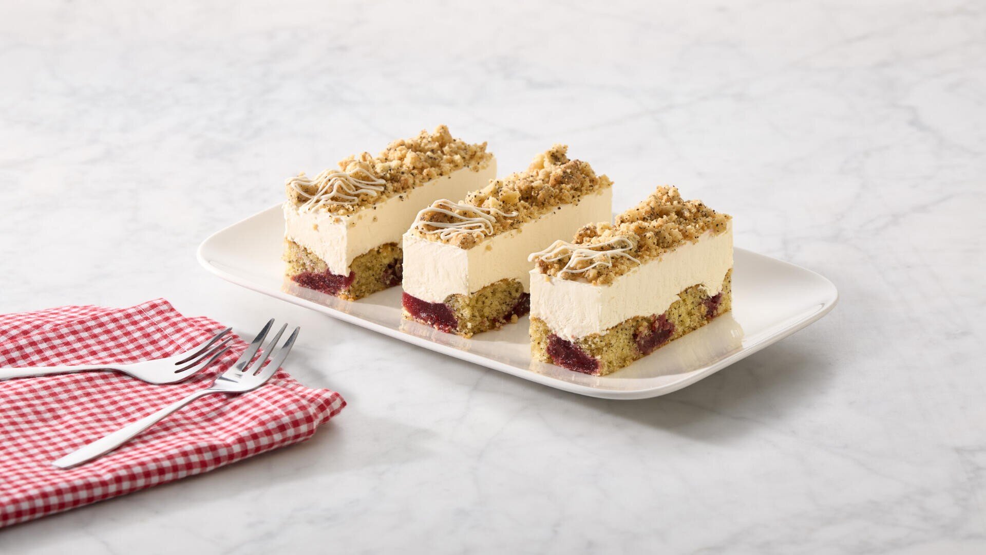 Drei rechteckige Scheiben des Schichtdesserts mit Sahne, Fruchtfüllung und Streuseln sind auf einem weißen Teller neben zwei Gabeln auf einer rot karierten Serviette auf einer Marmoroberfläche angerichtet.