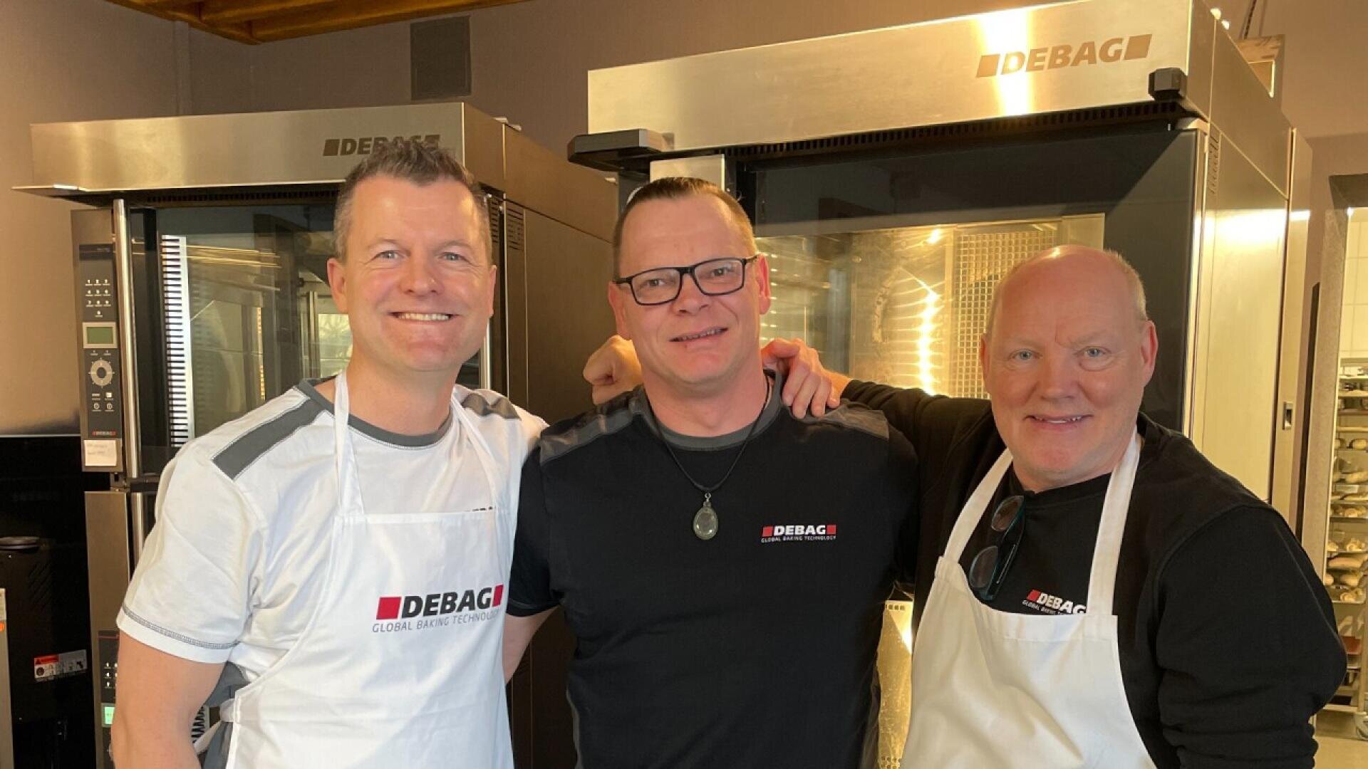Three men stand together smiling in a bakery kitchen, wearing DEBAG branded shirts and aprons, with two large commercial ovens in the background.