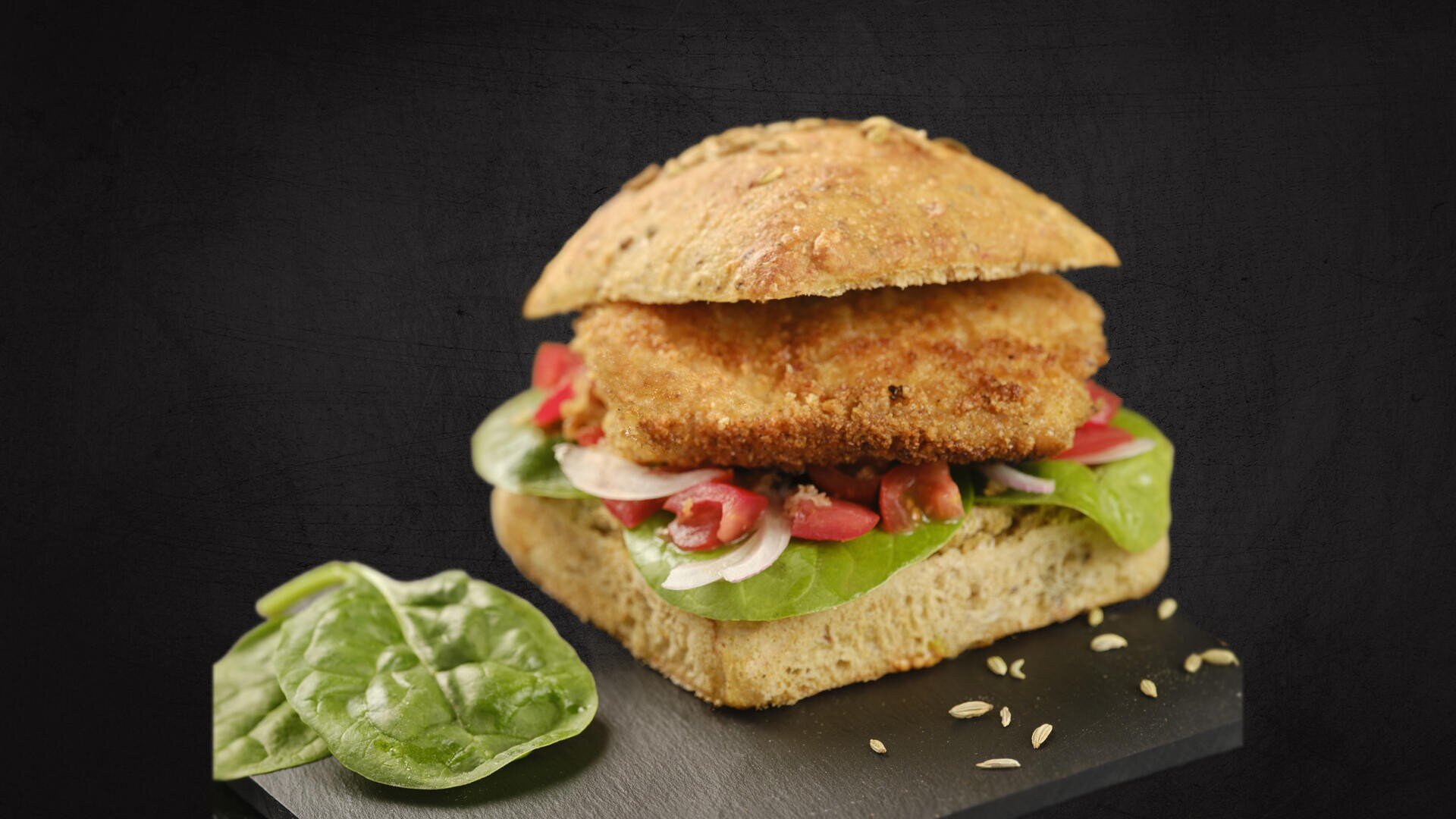 A sandwich with a square wholemeal roll, breaded patty, fresh spinach leaves, sliced tomatoes and onions. A spinach leaf and fennel seeds are placed on the slate in front of the sandwich.