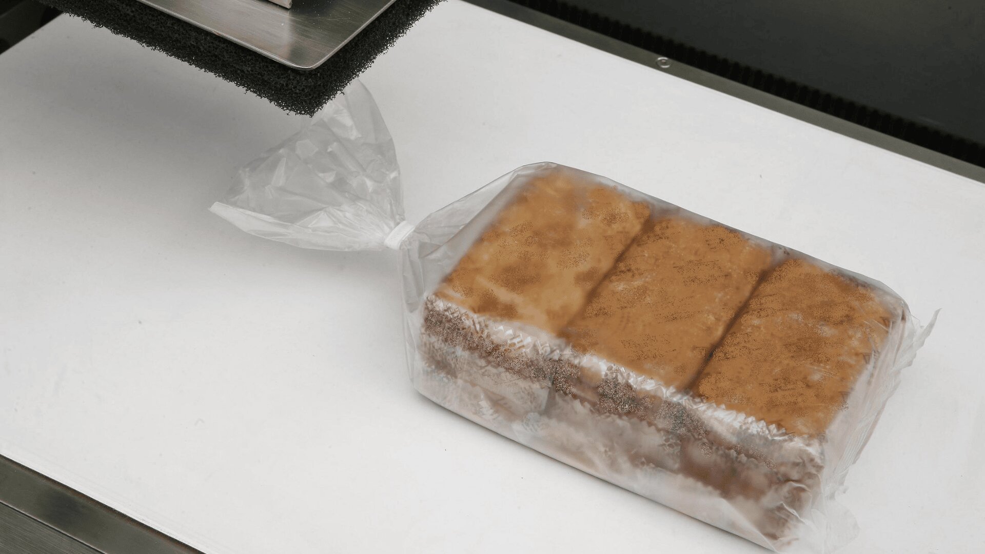 A loaf of bread, sliced and packaged in a clear plastic bag, sits on a white surface near a piece of industrial equipment.