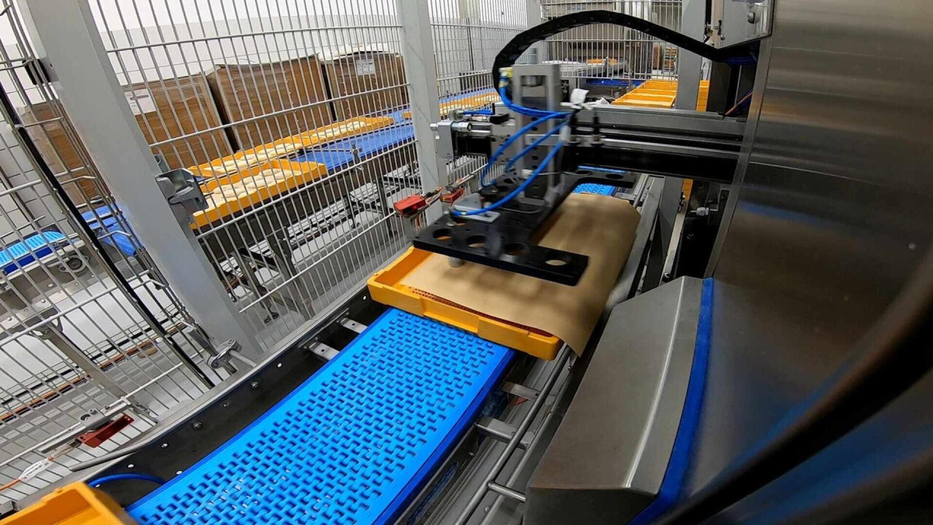 Ein Roboterarm legt in einer Industrieumgebung ein braunes Paket auf ein gelbes und blaues Förderband in einem umzäunten Bereich, in dem sich im Hintergrund Kartons stapeln.