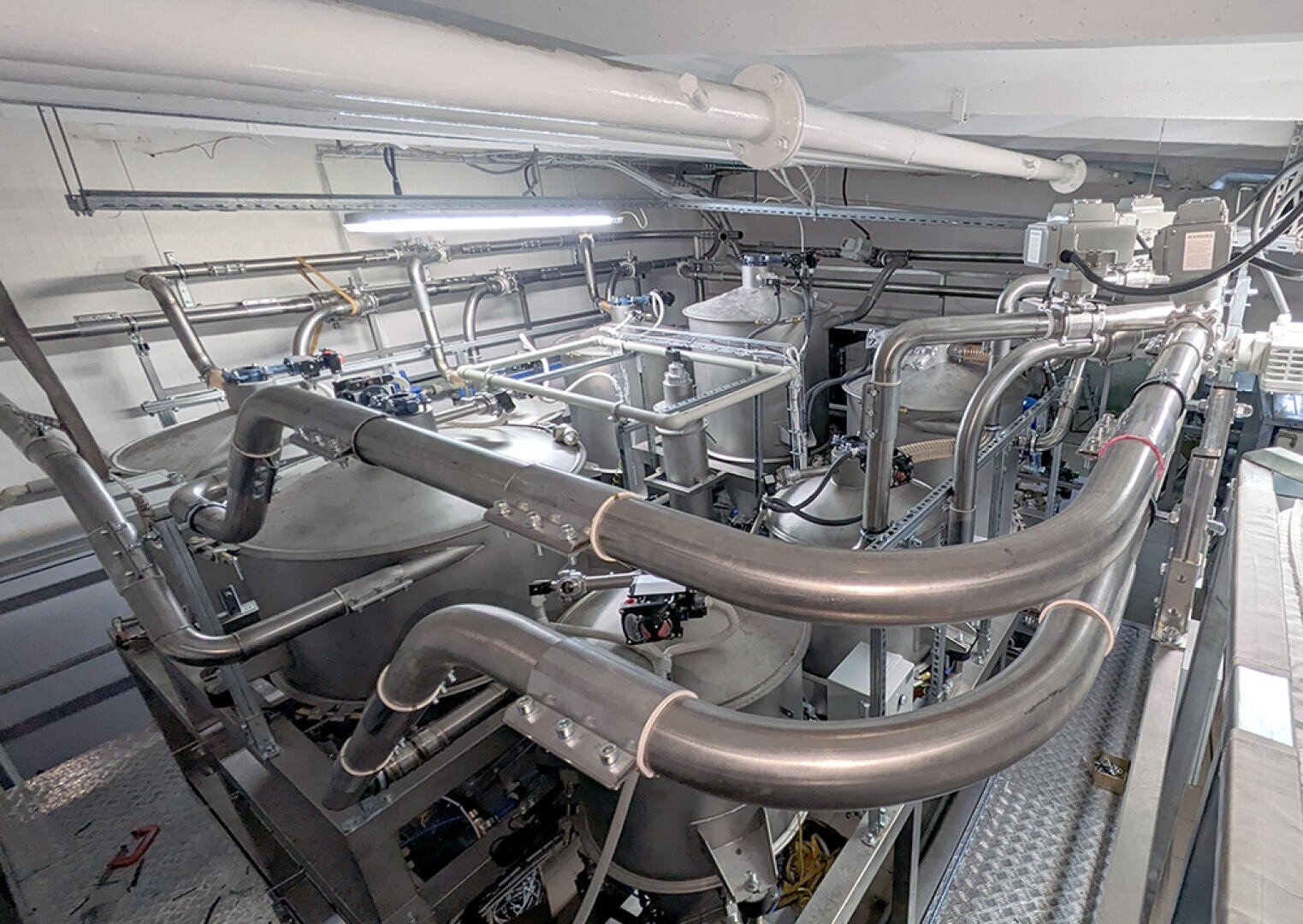 A complex industrial setup featuring large metal tanks, pipes, and valves connected throughout, likely part of a processing or manufacturing facility with overhead lighting illuminating the equipment.