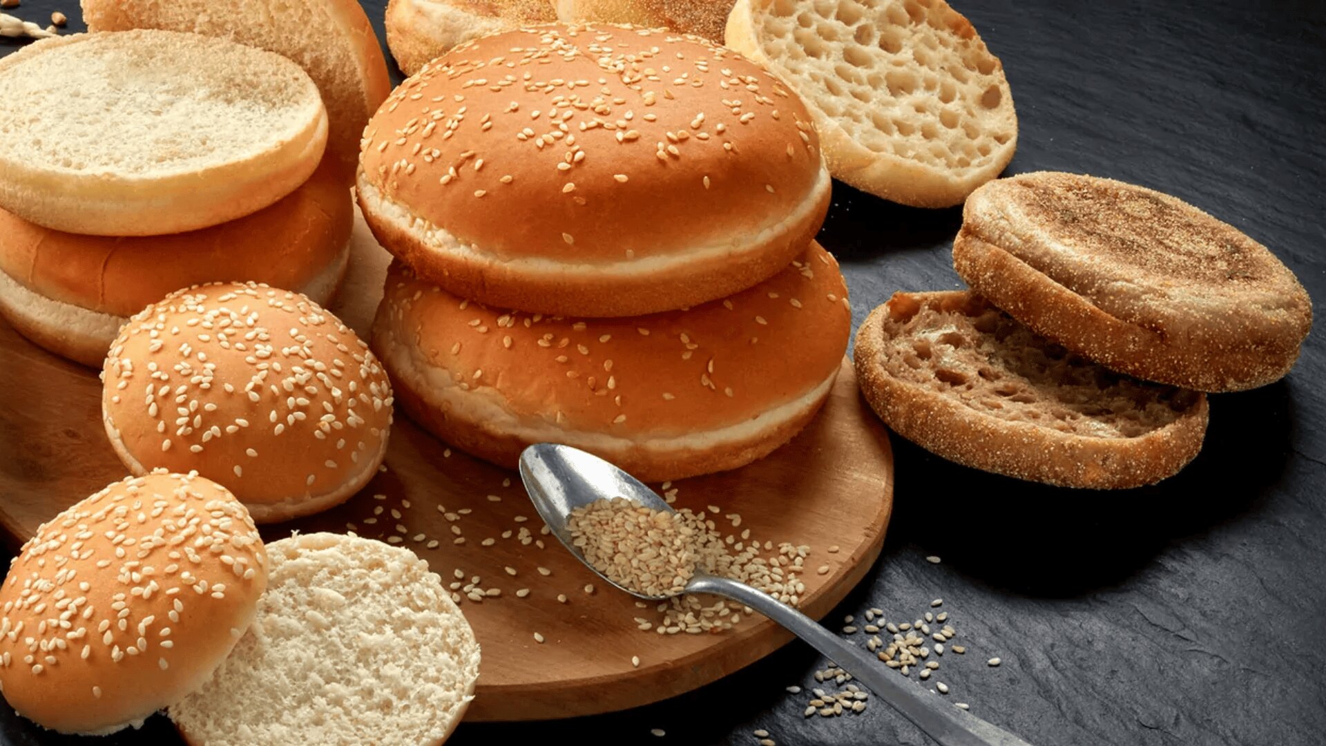 Auf einem Holzbrett und einer dunklen Fläche liegt eine Auswahl von Sesambrötchen und englischen Muffins, im Vordergrund ein Löffel mit Sesamkörnern.