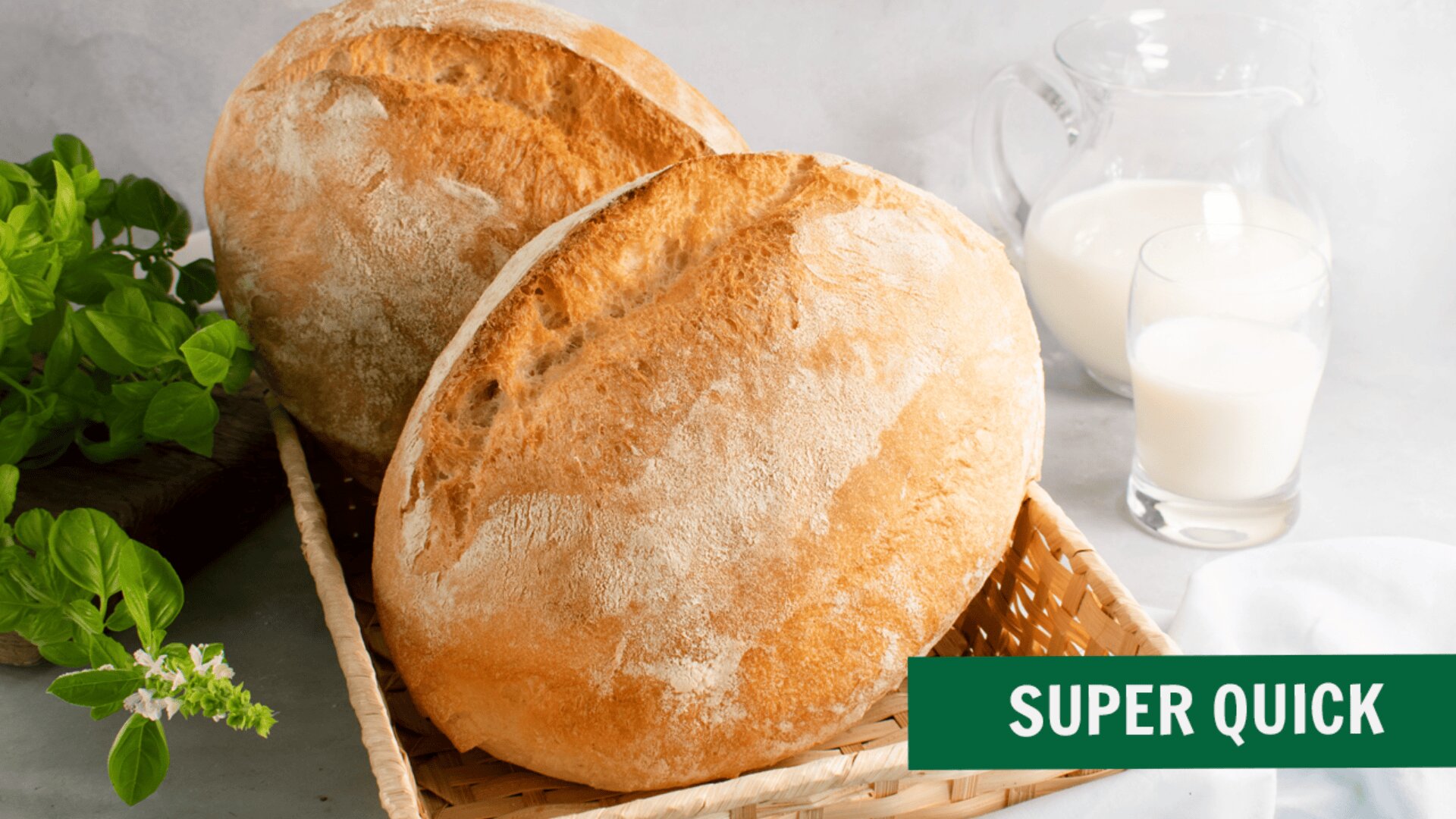 Two round loaves of crusty bread sit in a woven basket next to a green basil plant. In the background, a glass jug and a glass of milk are visible. A green label in the corner reads SUPER QUICK.