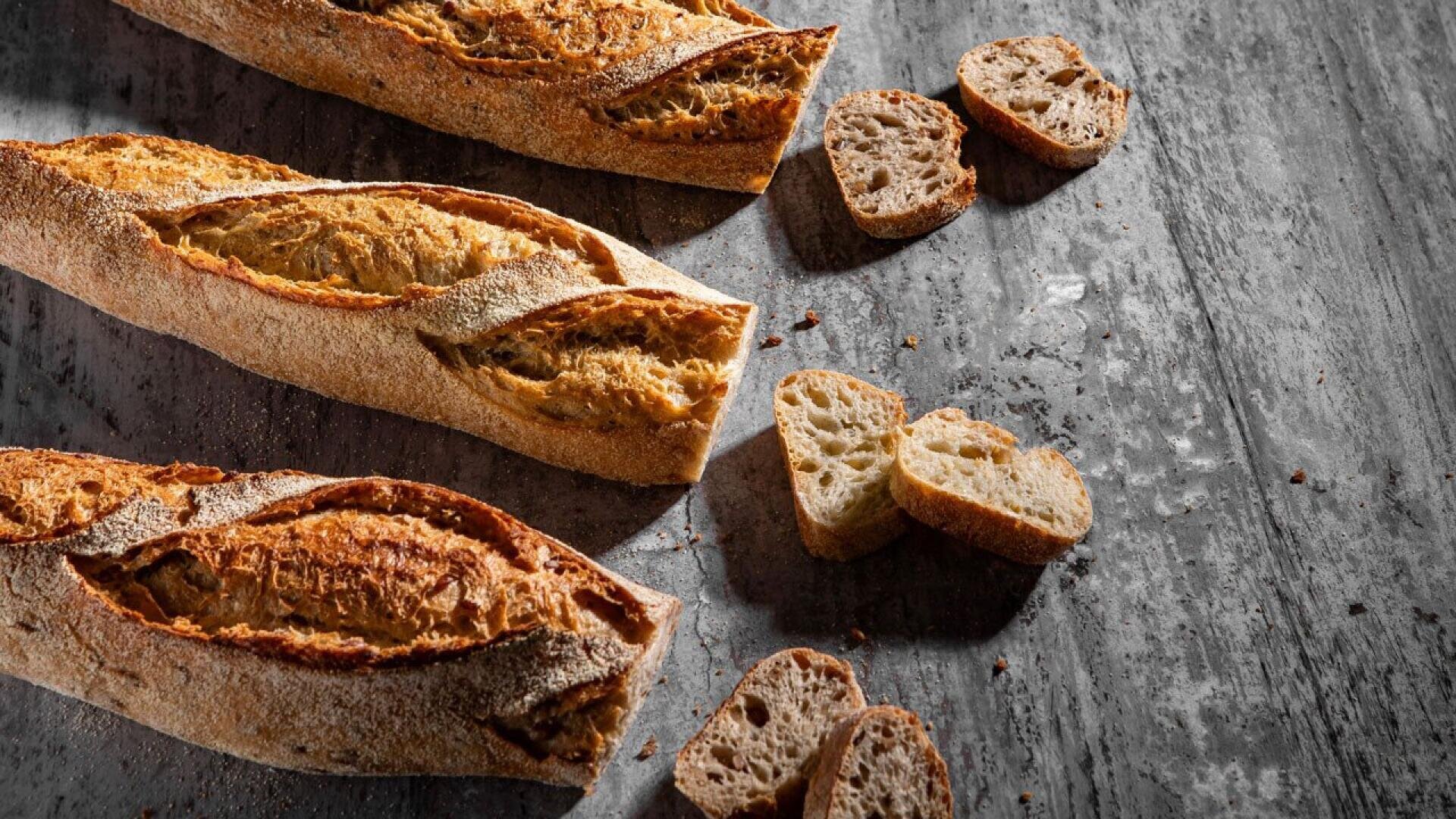 Three crusty baguettes and several slices of bread are arranged on a rustic wooden surface, with crumbs scattered around. The bread has a golden-brown, textured crust and airy interior.