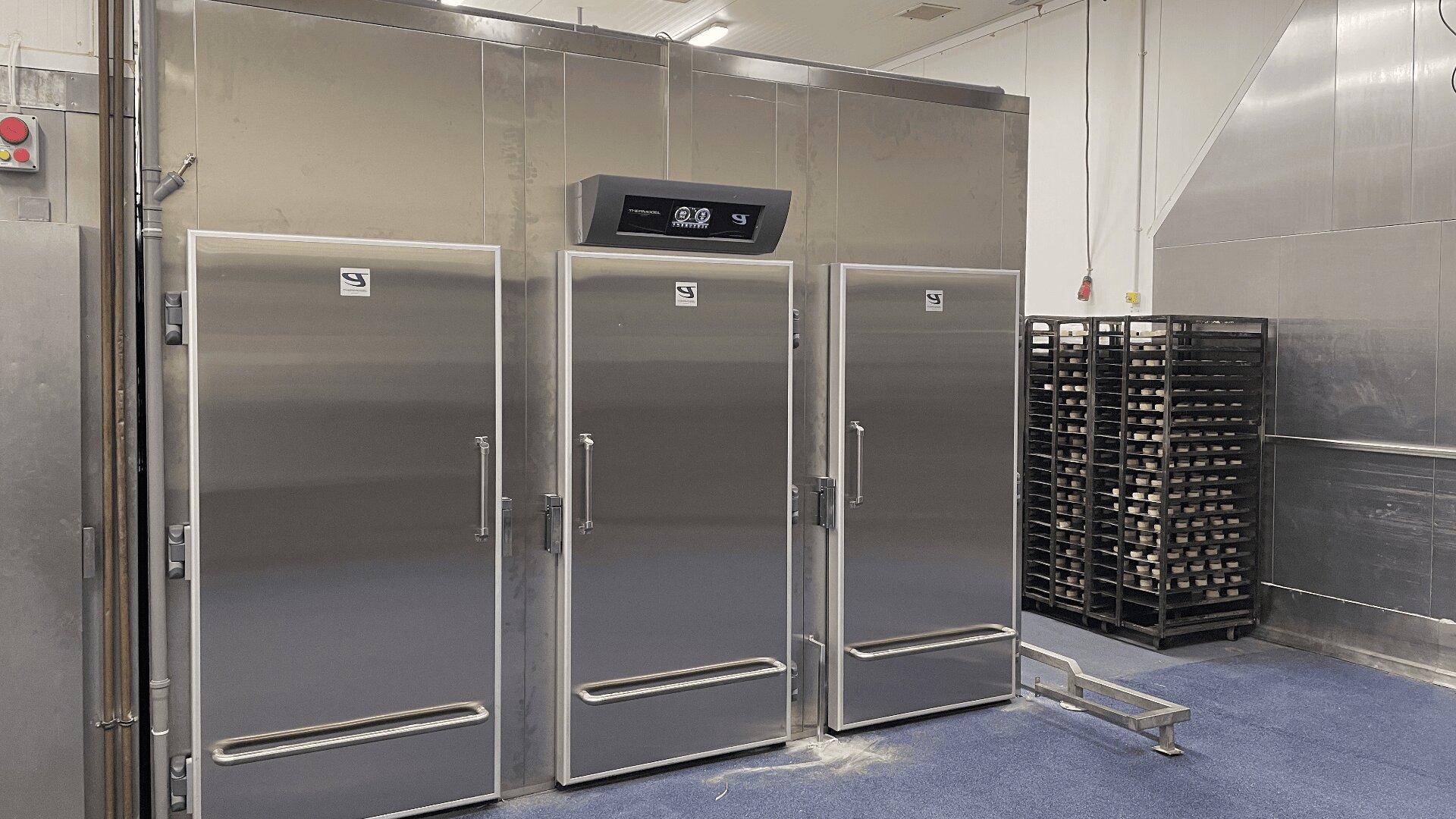 Three large stainless steel industrial refrigerators with digital displays are set against a clean wall in a commercial kitchen. To the right, metal racks hold trays. The floor is blue and the area is well-lit.