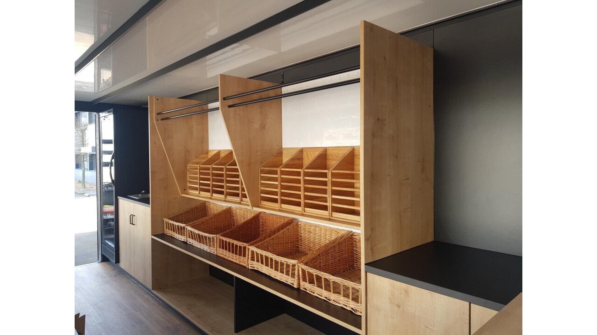 Wooden display shelves with wicker baskets and cubby holes, likely for bread or pastries, inside a modern, clean retail or market space. The area appears well-organized and ready for stocking.