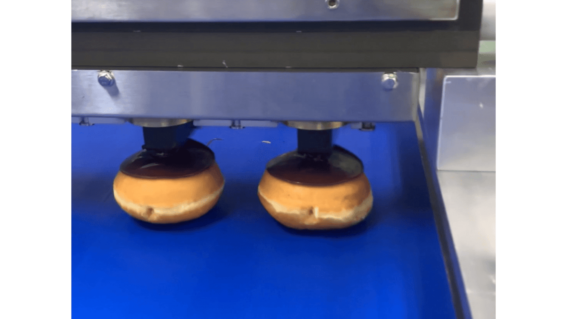 Zwei Krapfen auf einem Förderband werden von einer Maschine mit zwei runden Düsen mit Schokoladenglasur überzogen.