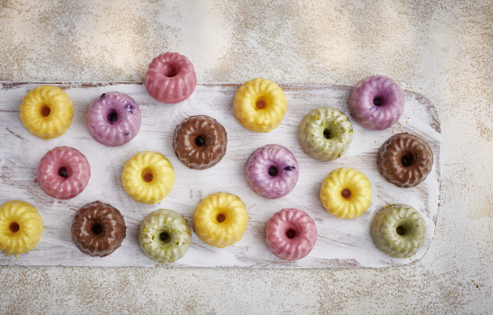 Colorful mini bundt cakes with glossy icing in pink, yellow, green, and brown are arranged in rows on a rustic white wooden board set on a light textured surface.