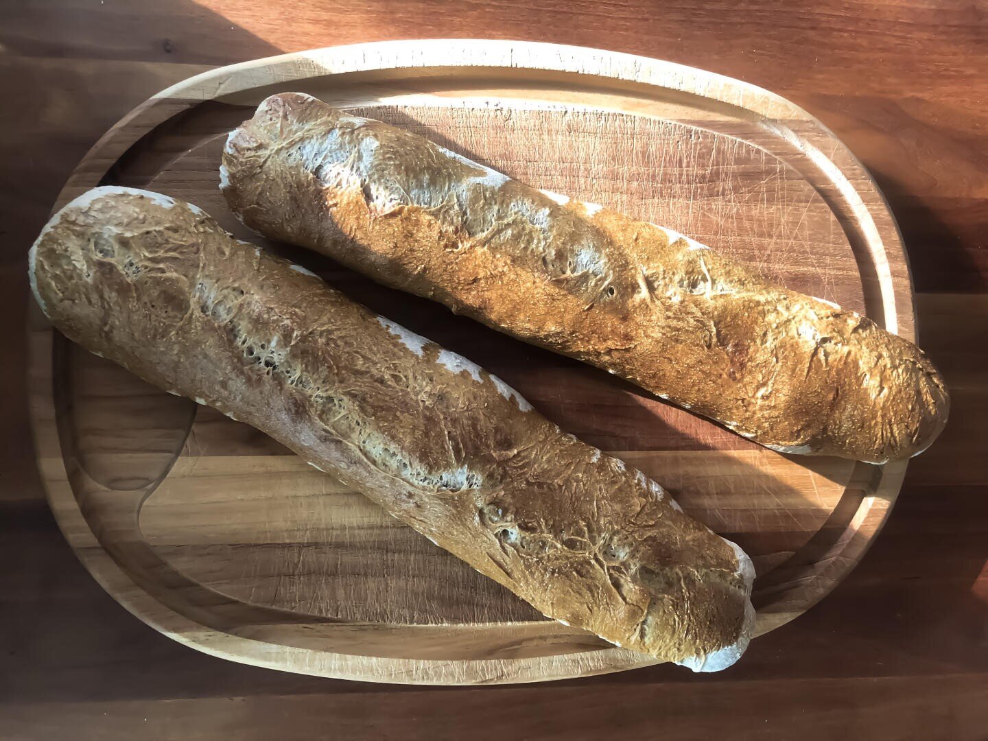 Two baguettes with golden, crusty tops rest side by side on a wooden cutting board, illuminated by sunlight.