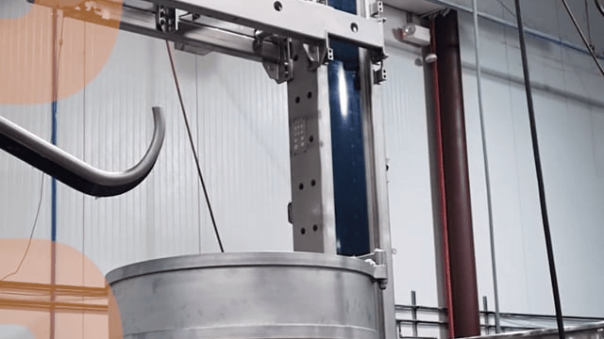 A large metal hook lifts a cylindrical metal container inside an industrial facility with white paneled walls and exposed pipes.