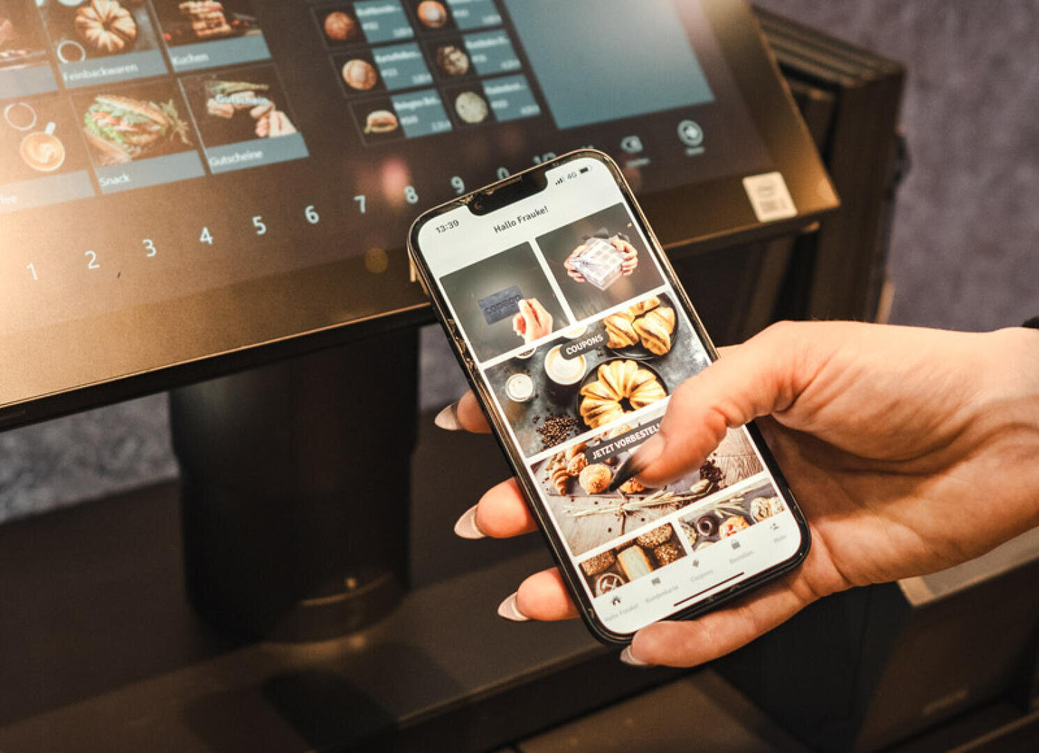 Eine Person hält ein Smartphone mit einer App zur Essensbestellung und zur Auswahl von Backwaren vor einem Touchscreen-Kiosk mit einer Speisekarte.