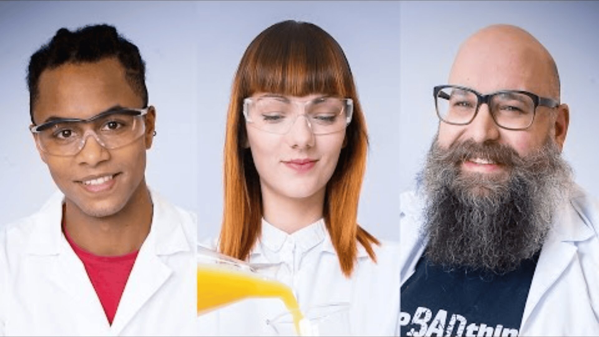 Drei Personen in Laborkitteln und mit Schutzbrillen stehen nebeneinander. Der erste lächelt in die Kamera, der zweite gießt eine gelbe Flüssigkeit ein, und der dritte, mit einem langen Bart und einer Brille, lächelt ebenfalls fröhlich.