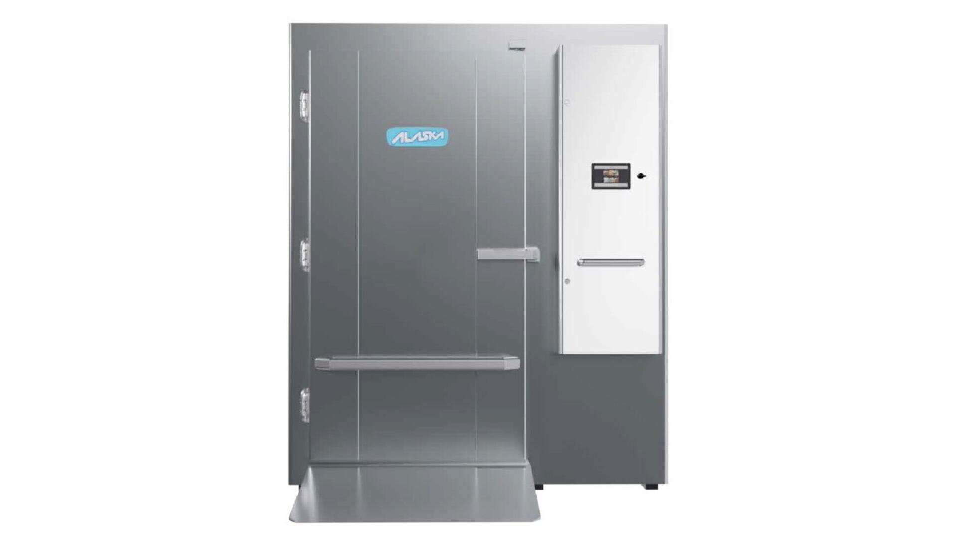 Ein großer, grauer, begehbarer Industriekühl- oder -gefrierschrank mit einem Türgriff, einer Stufenrampe und einem Bedienfeld auf der rechten Seite, das das ALASKA-Logo auf der Vorderseite trägt.