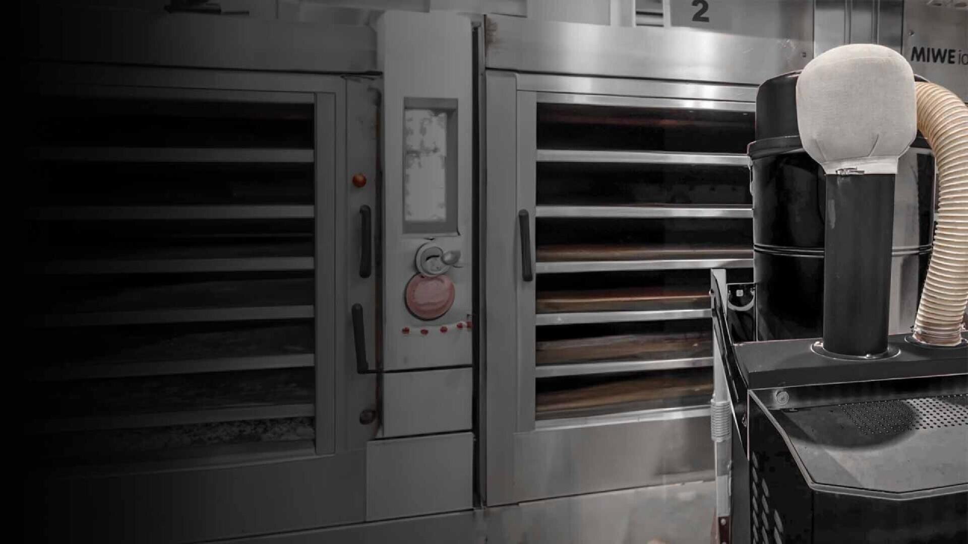 A commercial bakery oven with several shelves is shown next to a large black machine with a beige hose and a white cylindrical attachment in a modern industrial kitchen.