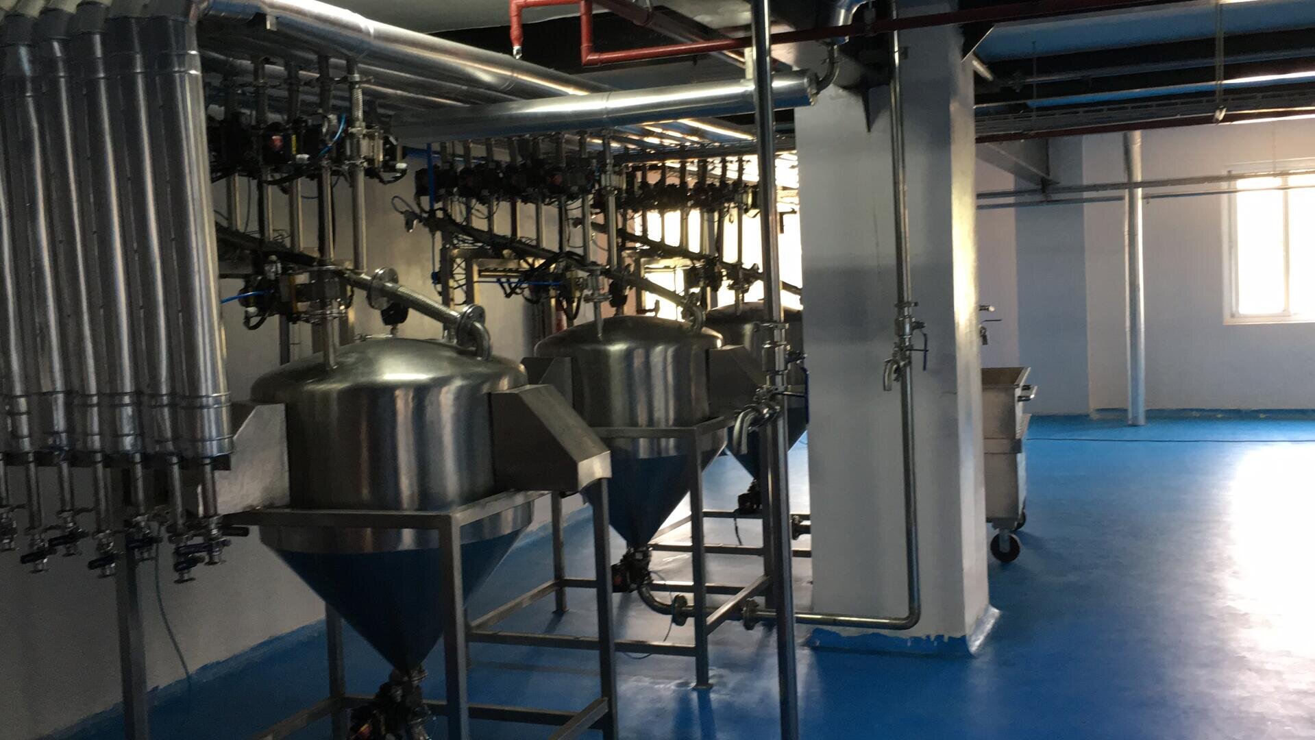 Large stainless steel industrial tanks and pipes in a clean, blue-floored factory setting. The tanks have cone-shaped bottoms, and various control valves and metal pipes are visible overhead. Light enters from a window.