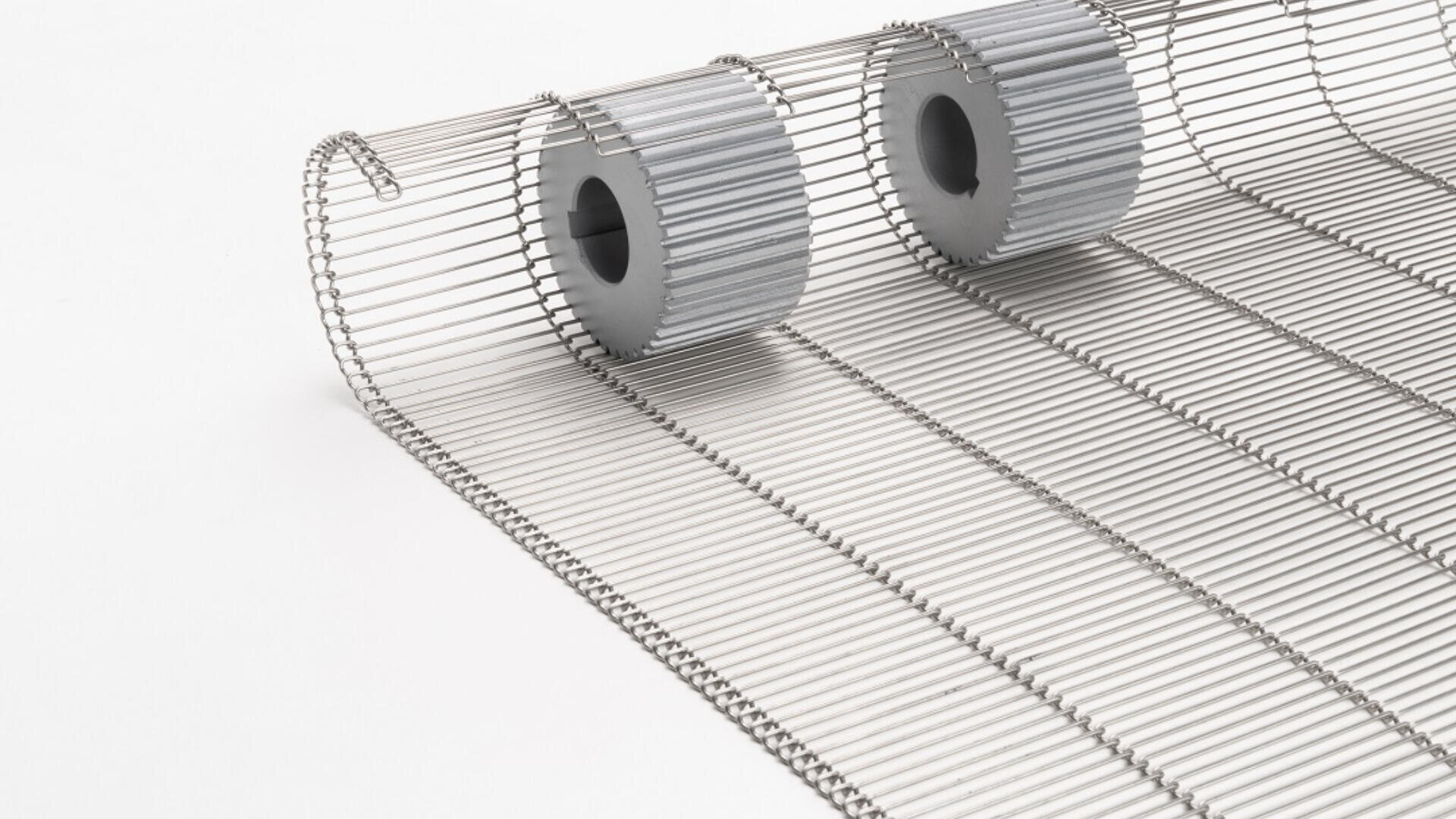 Close-up of a metallic wire conveyor belt partially unrolled, with two grey metal sprockets placed on top of the belt, all set against a white background.