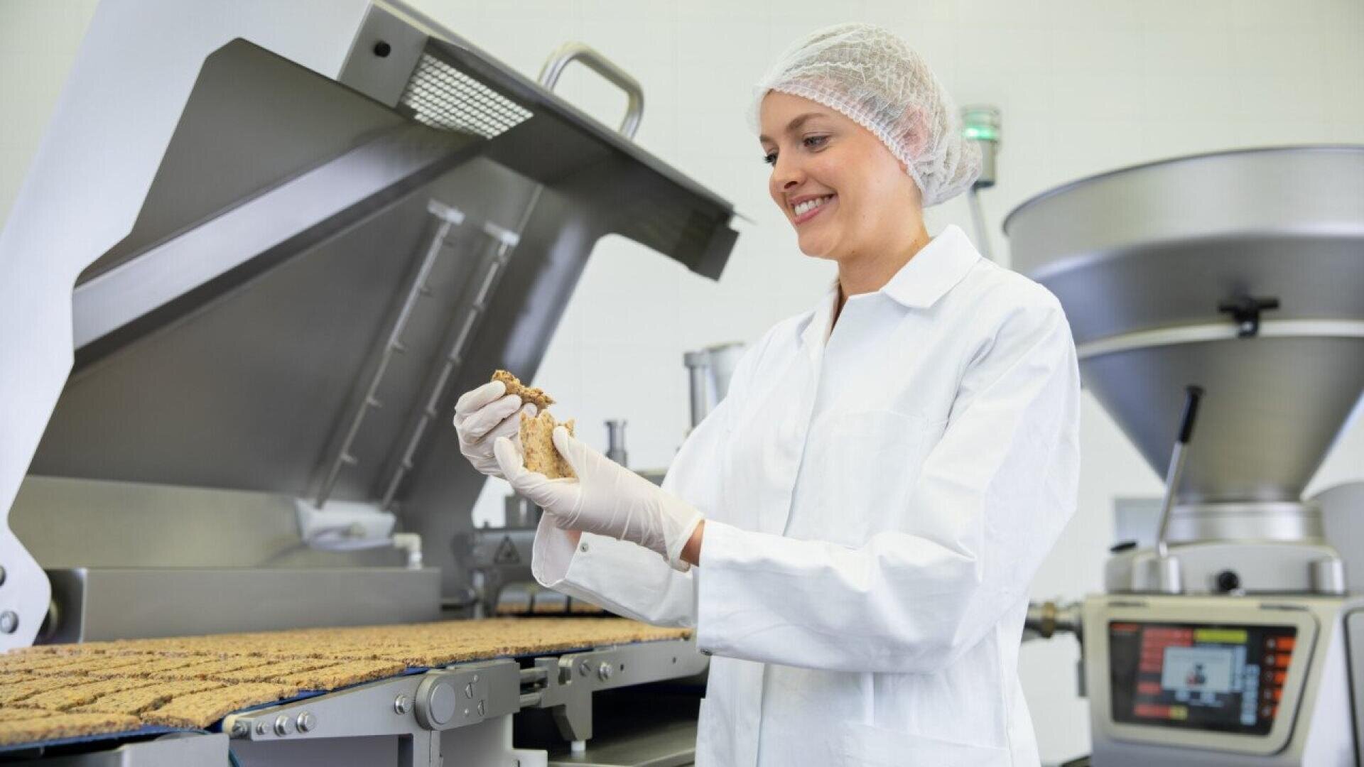 A woman in a lab coat, hairnet, and gloves inspects a food product on a production line in a factory setting, surrounded by industrial food processing equipment.