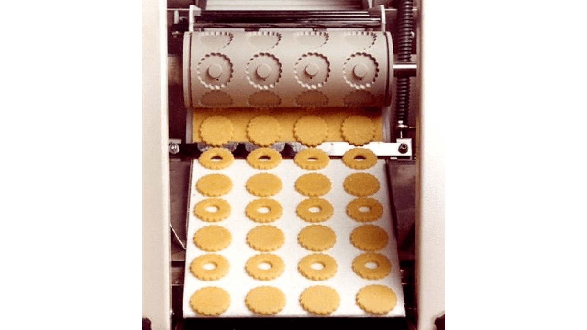 A machine presses dough, cutting out uniform round cookies with fluted edges and central holes, and places them in neat rows on a conveyor belt.