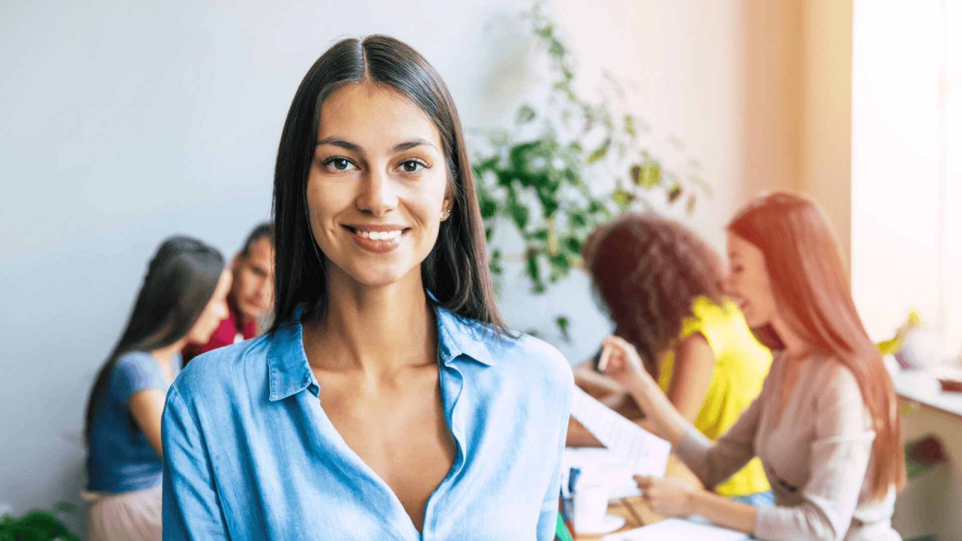 A woman in a blue shirt smiles at the camera, standing in a bright office space. In the background, four women are engaged in conversation and work around a table, with a plant visible by the window.
