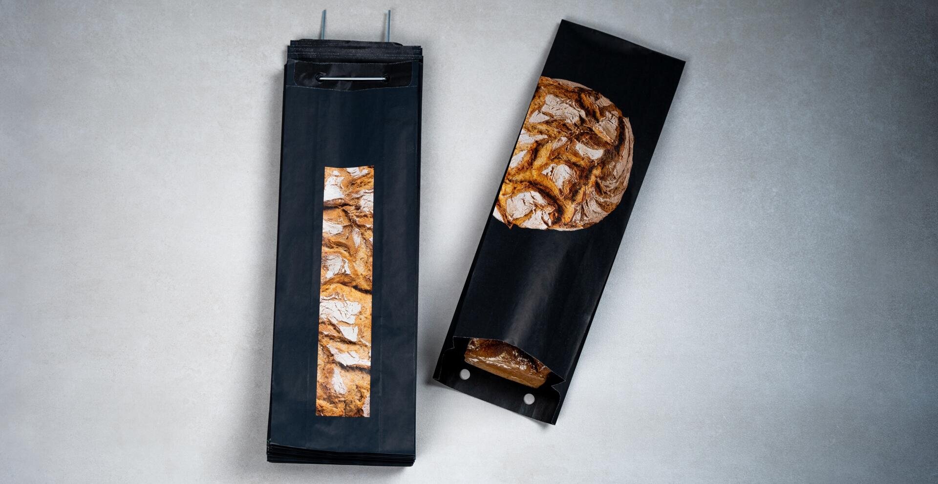 Zwei schwarze Brottüten aus Papier mit einem durchsichtigen Fenster und einem aufgedruckten Bild von rustikalem Brot; eine Tüte ist mit anderen gestapelt und die zweite Tüte enthält einen Laib Brot, der teilweise durch das Fenster sichtbar ist.