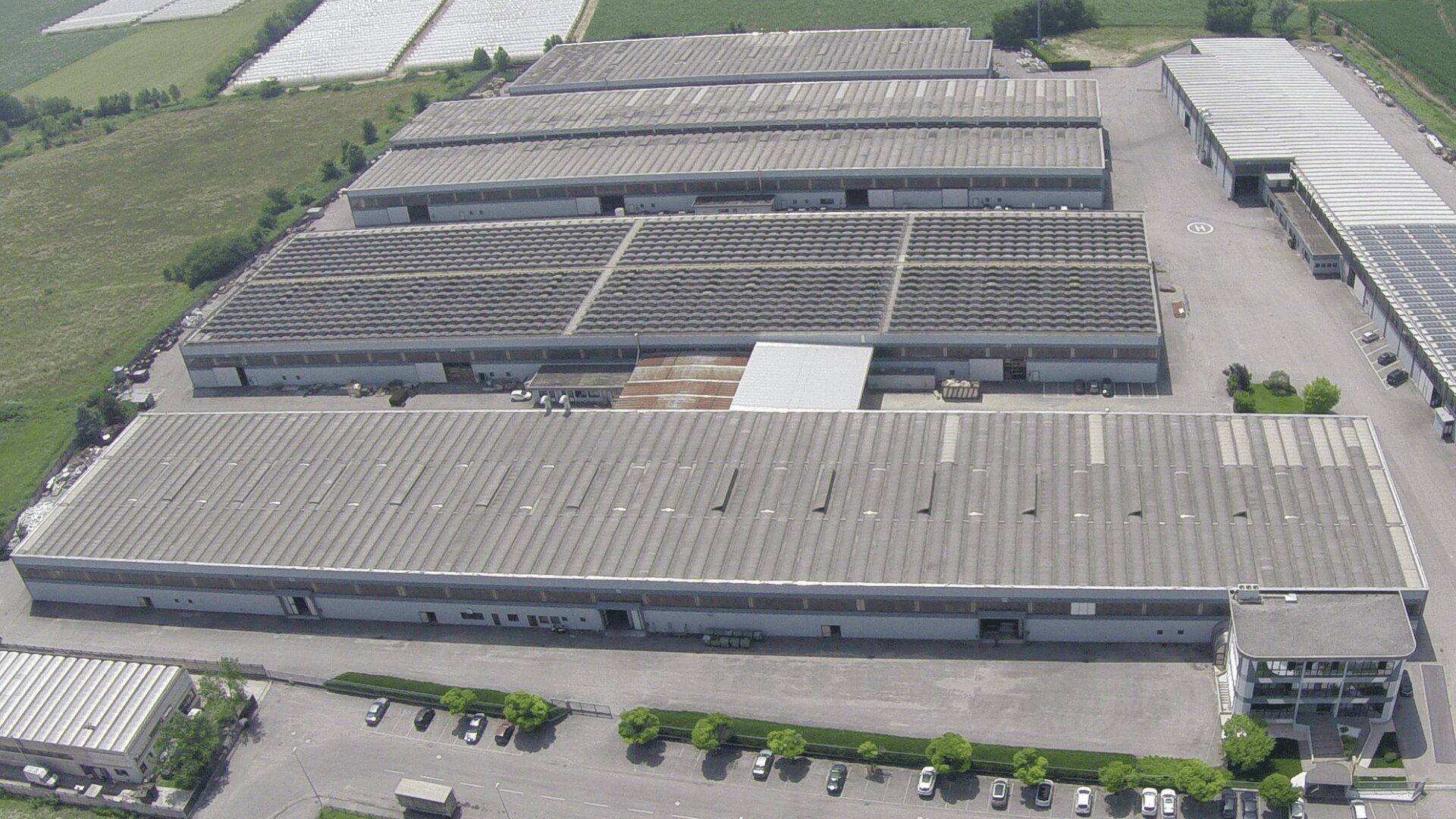 Aerial view of a large industrial warehouse complex with multiple long, rectangular buildings, gray roofs, loading docks, and parking areas surrounded by greenery and fields.