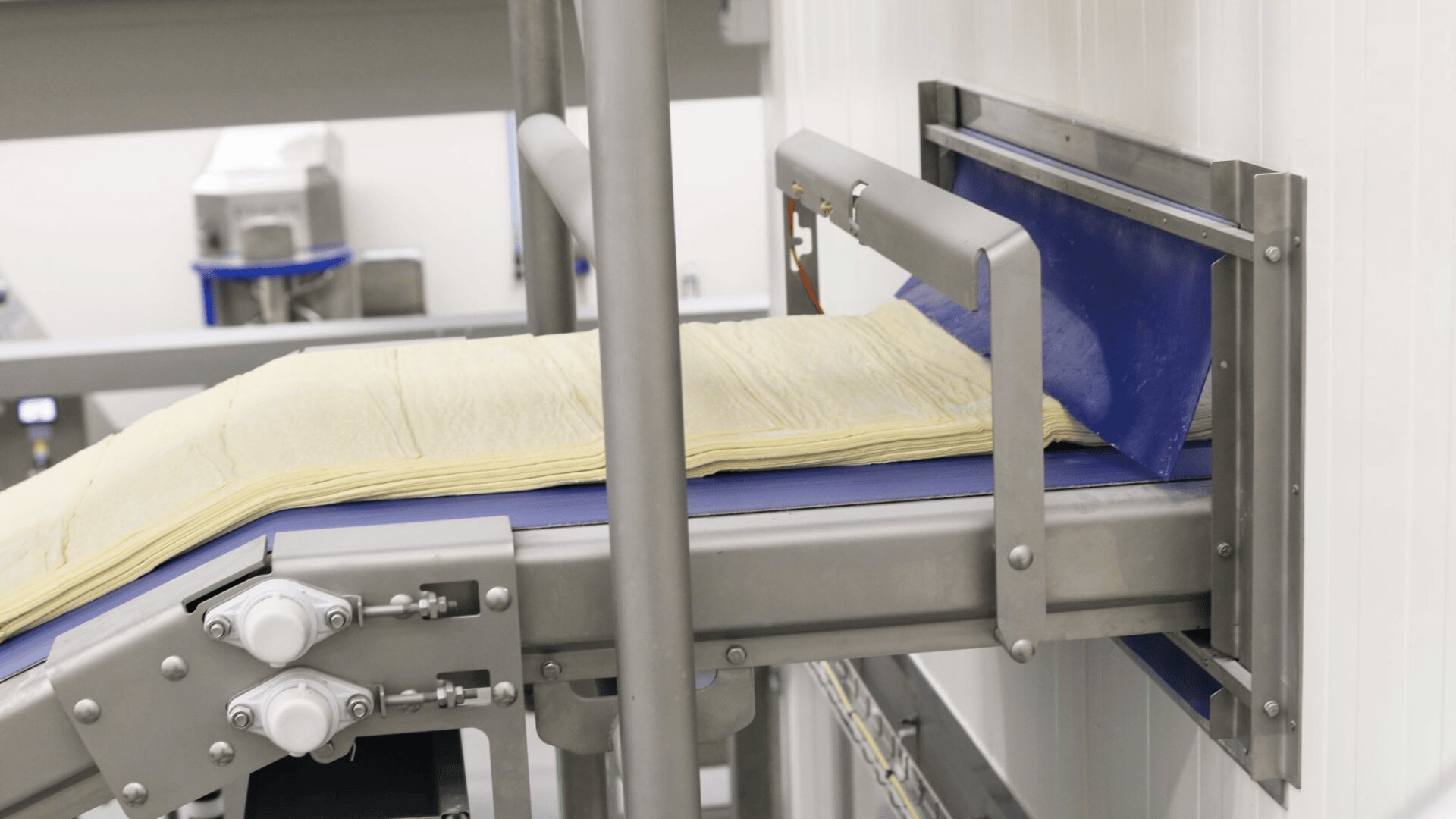 Ein Förderband transportiert Teigschichten durch eine Öffnung in einer Metallwand in einer industriellen Lebensmittelverarbeitungsanlage.