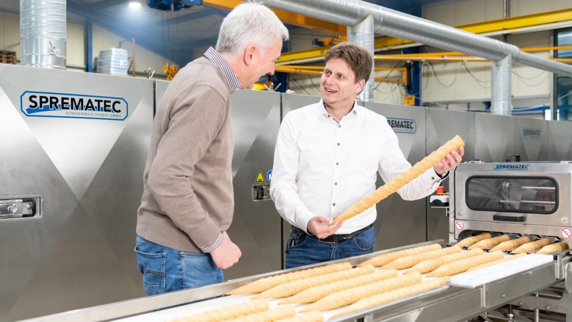 Two men stand beside a conveyor belt with long, rolled pastry dough in a factory setting, discussing or inspecting one of the pastries. Industrial baking machines labeled SPREMATEC are visible in the background.