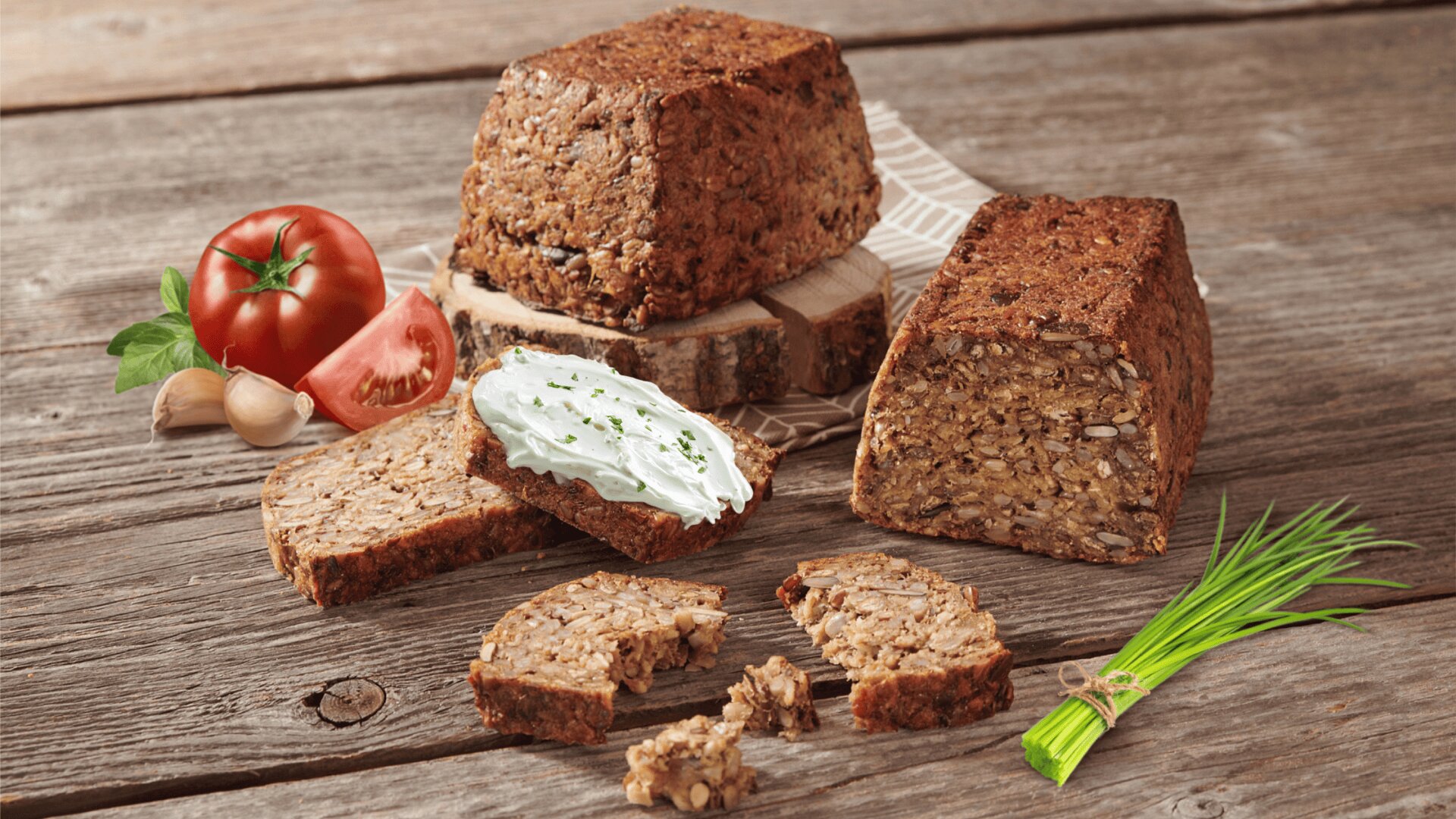 A rustic wooden table with seed bread loaves and slices, one topped with cream cheese and chives, beside a tomato, garlic cloves, and a bunch of fresh chives.