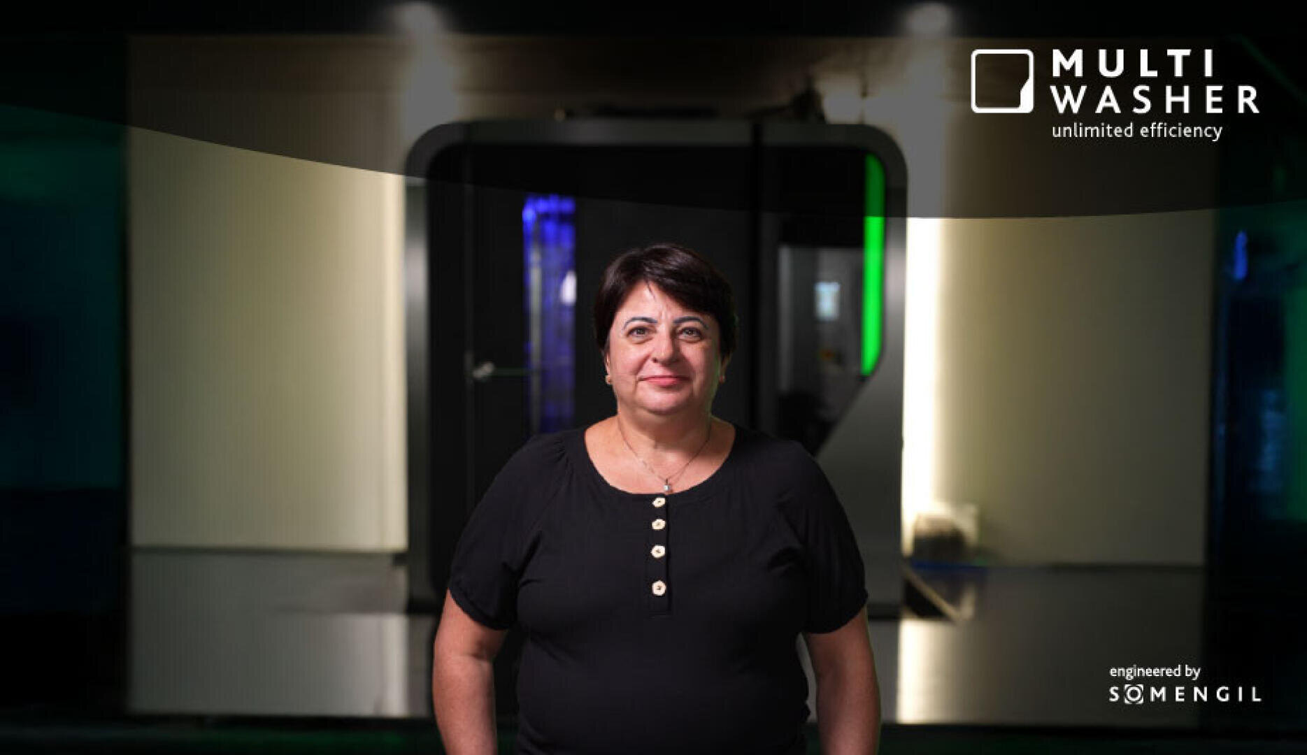 Eine Frau mit kurzen dunklen Haaren und einem schwarzen Hemd steht vor einer modernen Industriewaschmaschine. In der oberen rechten Ecke steht der Text „MULTI WASHER unlimited efficiency and engineered by SOMENGIL“.