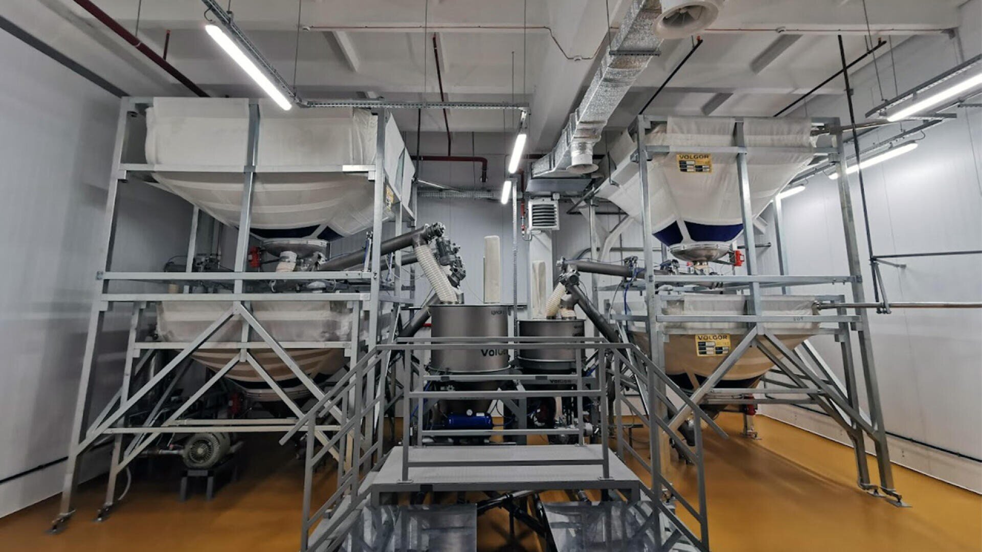 Large industrial machinery with metal frames and fabric-covered hoppers in a clean, well-lit factory room with yellow flooring and exposed pipes on the ceiling.