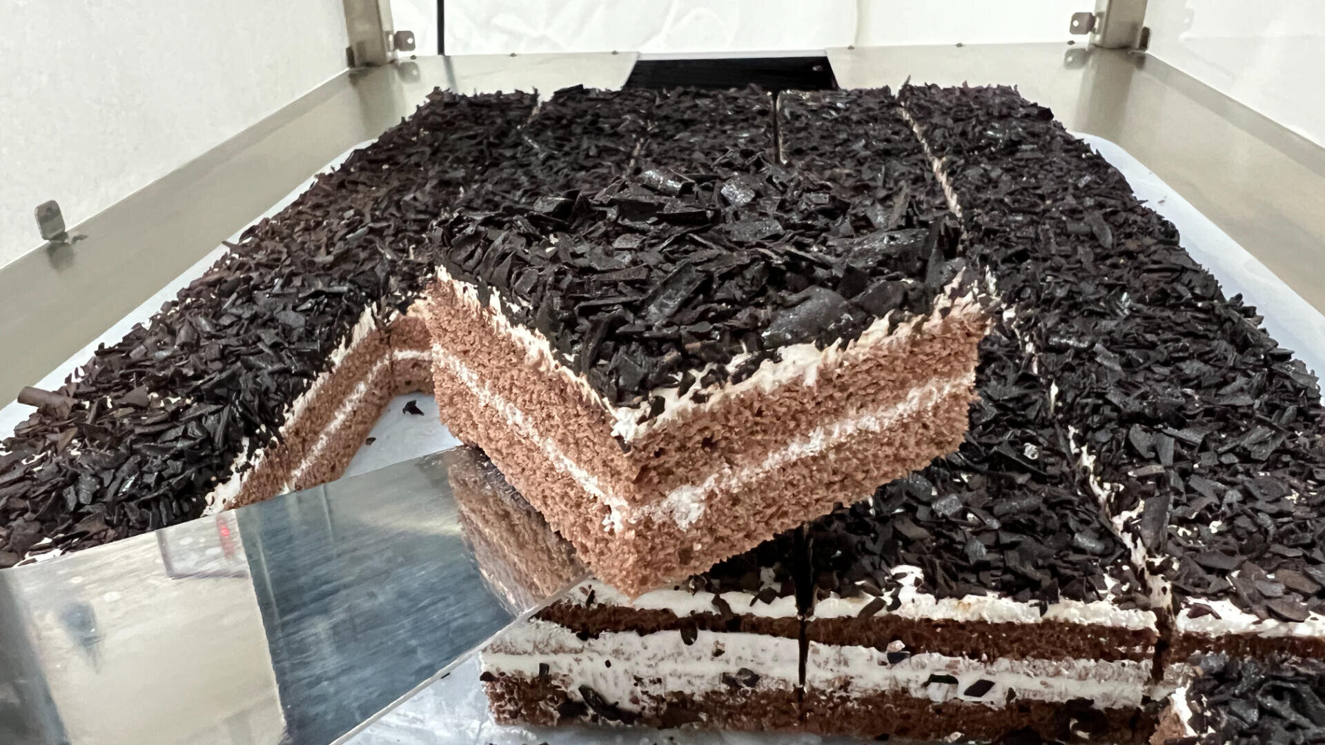A large rectangular chocolate cake topped with chocolate shavings is being sliced, with one piece lifted on a spatula, showing layers of chocolate and cream filling.