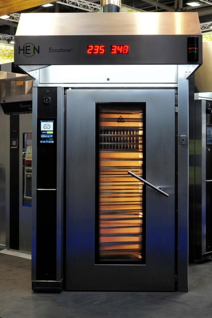Ein großer handelsüblicher Backofen aus Edelstahl mit einer Glastür, in der mehrere Backbleche zu sehen sind. Eine Digitalanzeige über der Tür zeigt die Zahlen 235 und 348 in Rot an. Der Ofen ist mit HEN Ecostone beschriftet.