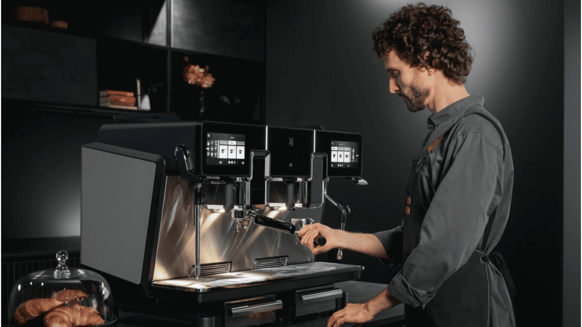 A barista operates a modern, dual-group espresso machine in a sleek, dark café setting. He wears a dark apron and shirt, with pastries visible on the counter nearby.