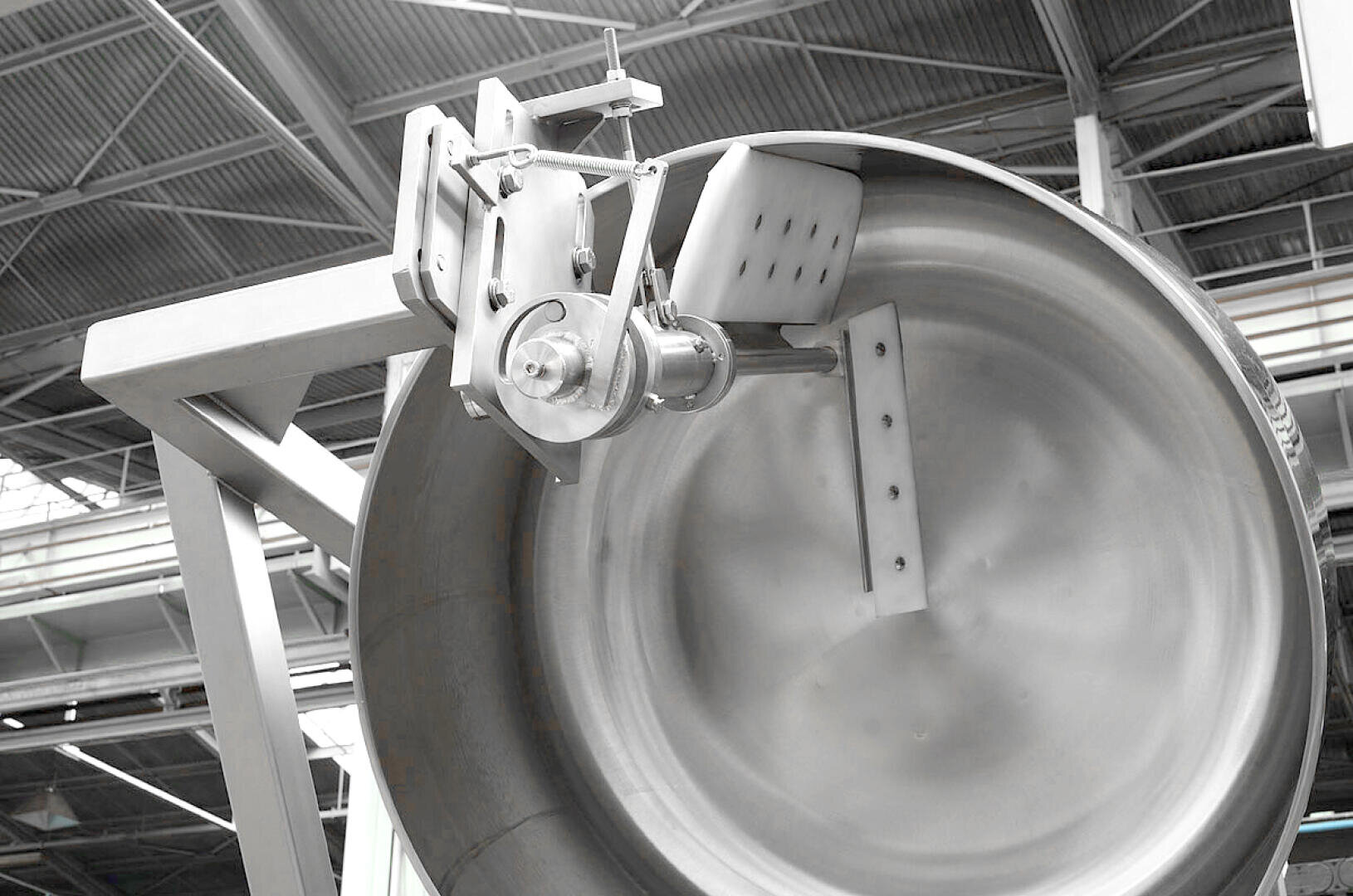 Large industrial stainless steel mixing drum with mechanical arm and paddle, inside a factory with high ceilings and metal beams visible in the background.